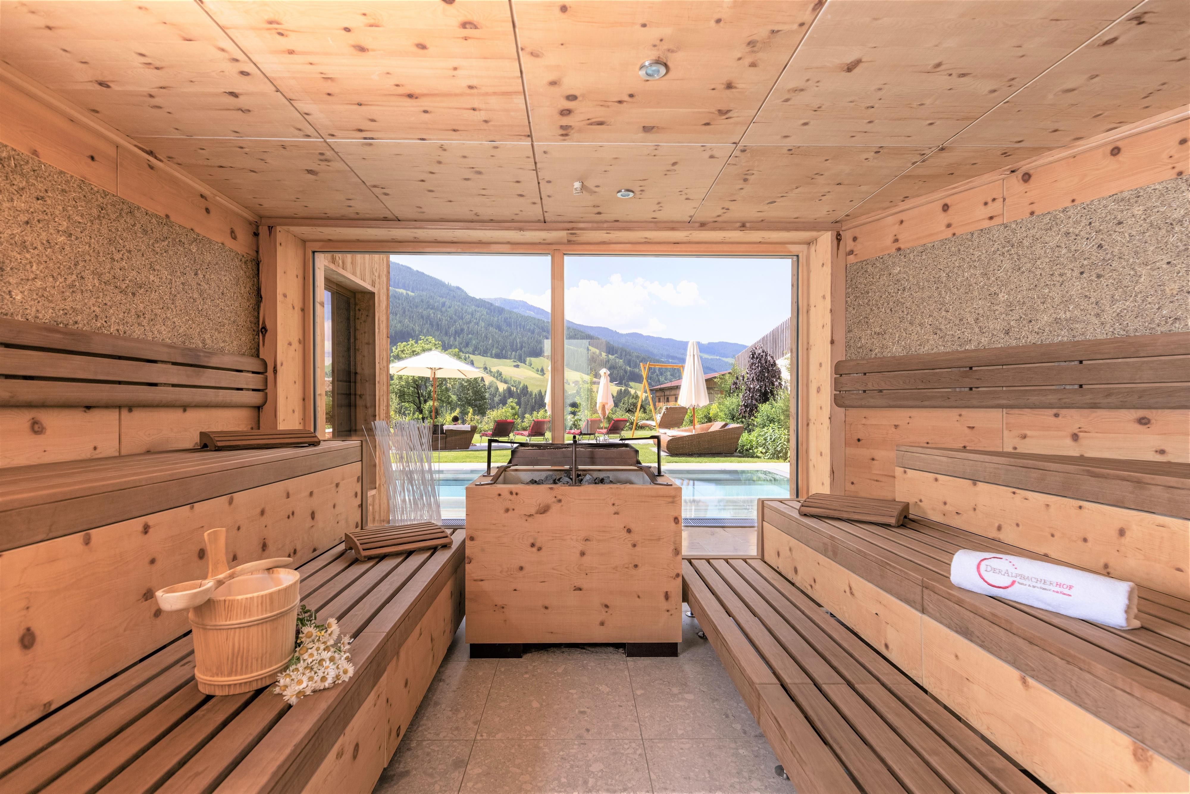 A cozy sauna area with wooden benches and a large window. The view shows a green landscape with an outdoor swimming pool.