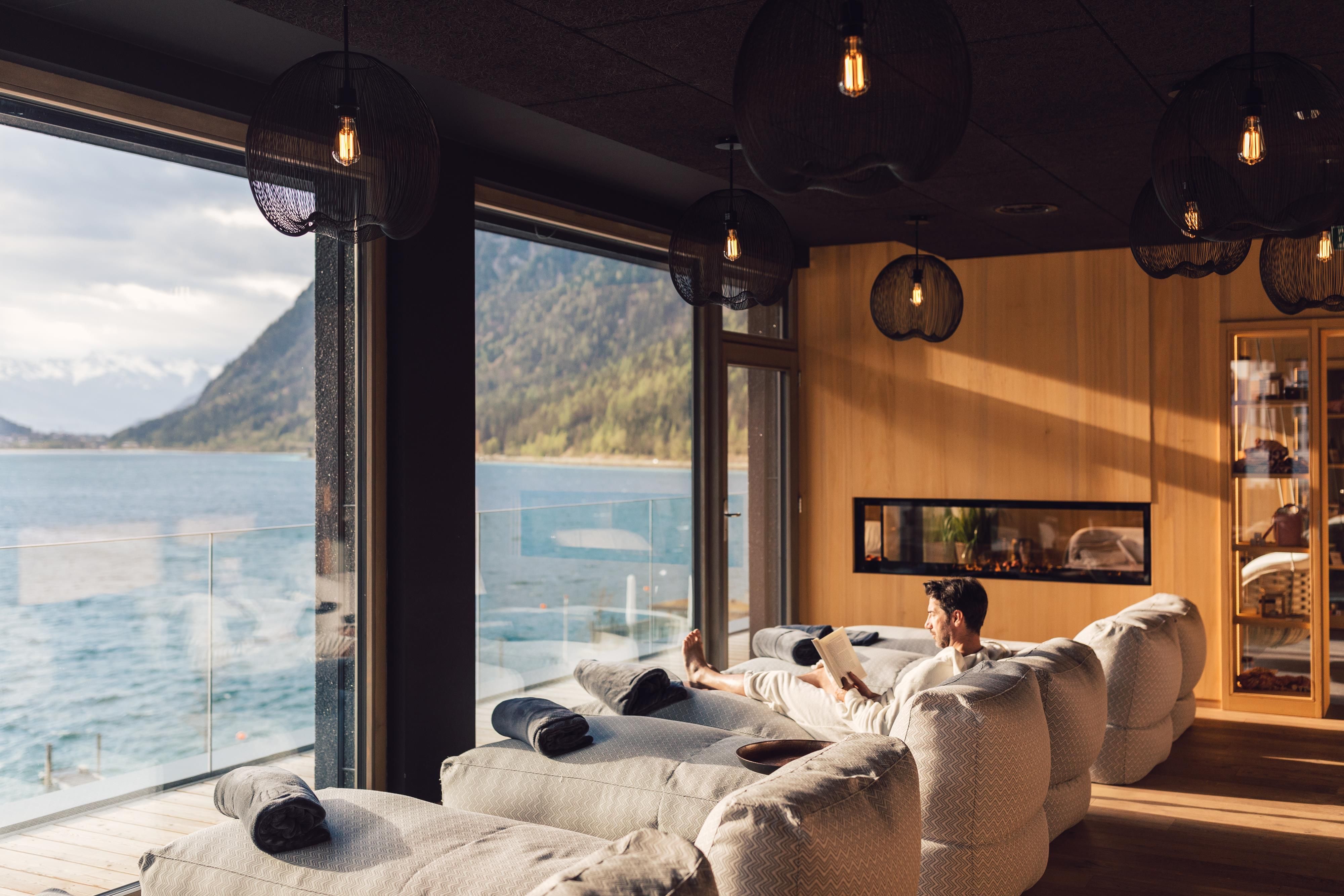 A cozy room with large windows and a view of a lake. A person is relaxing on a comfortable lounge chair.