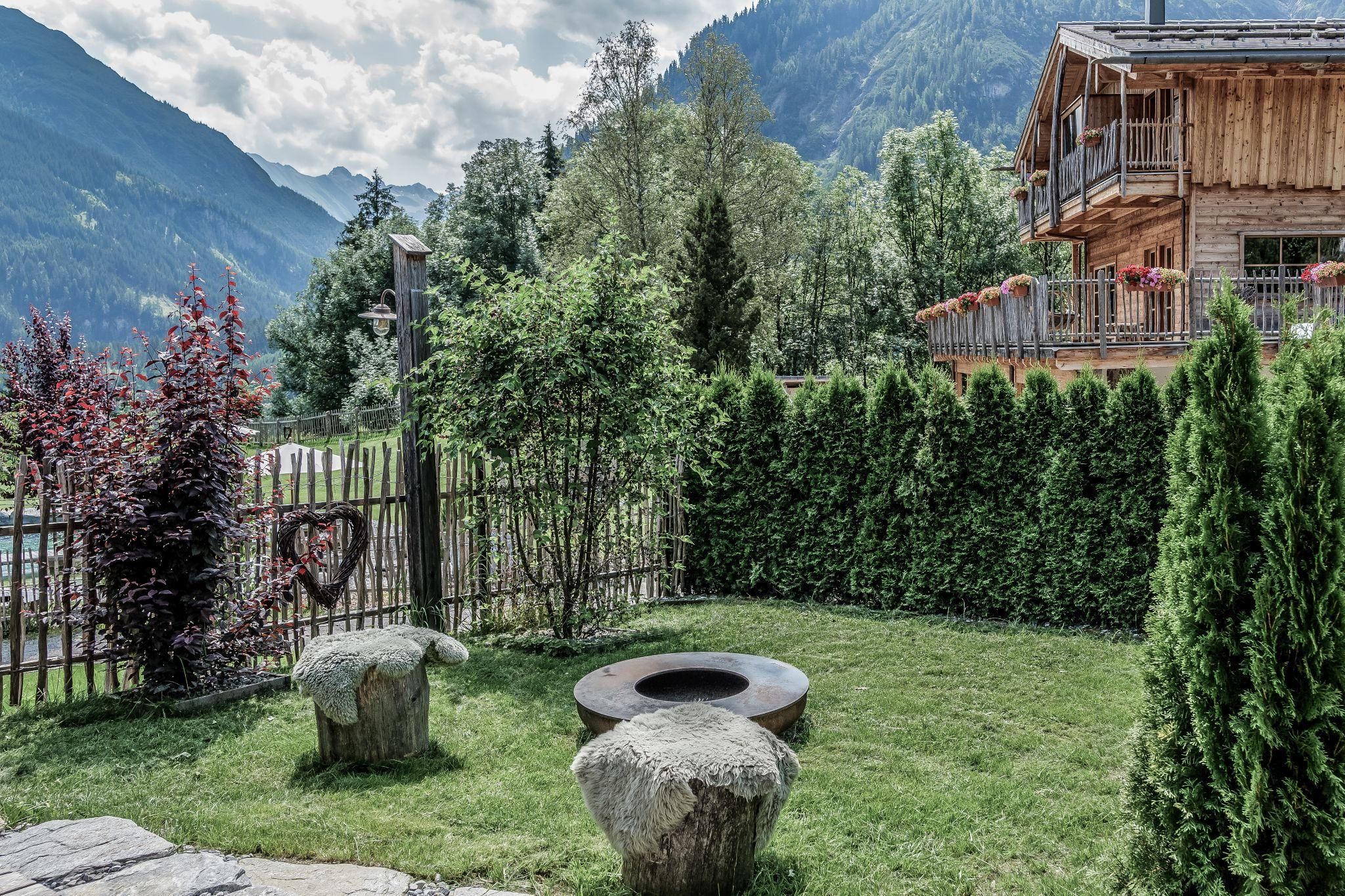A beautiful garden with green plants and a small stone table. In the background, there is a chalet and mountains visible.