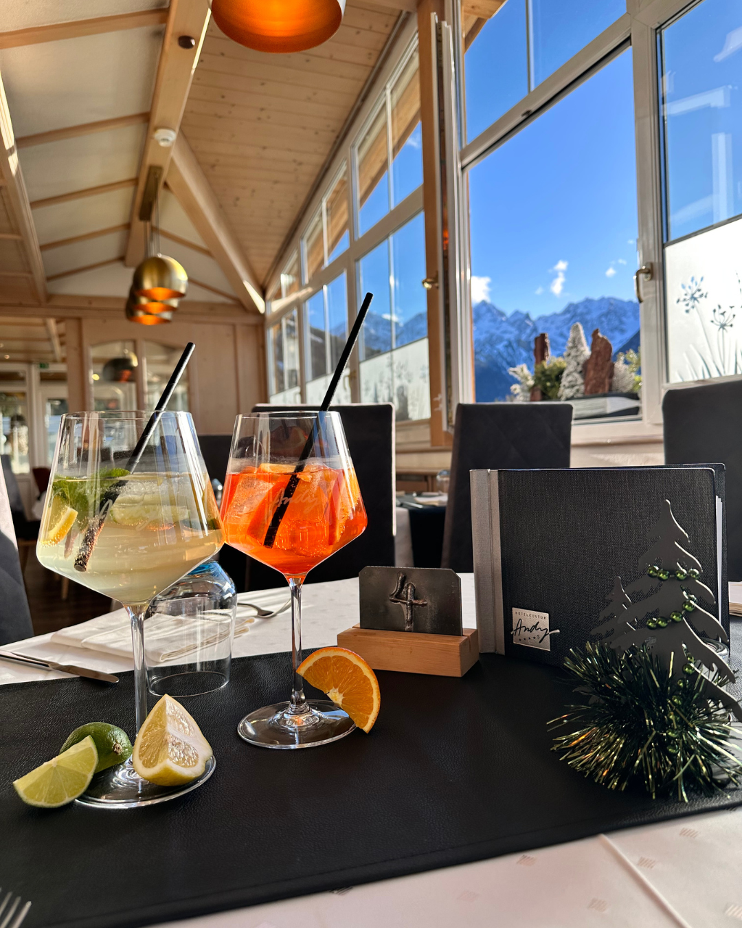 Two cocktails on a table in a restaurant with large windows and a mountain view. The room is bright and cozily furnished.