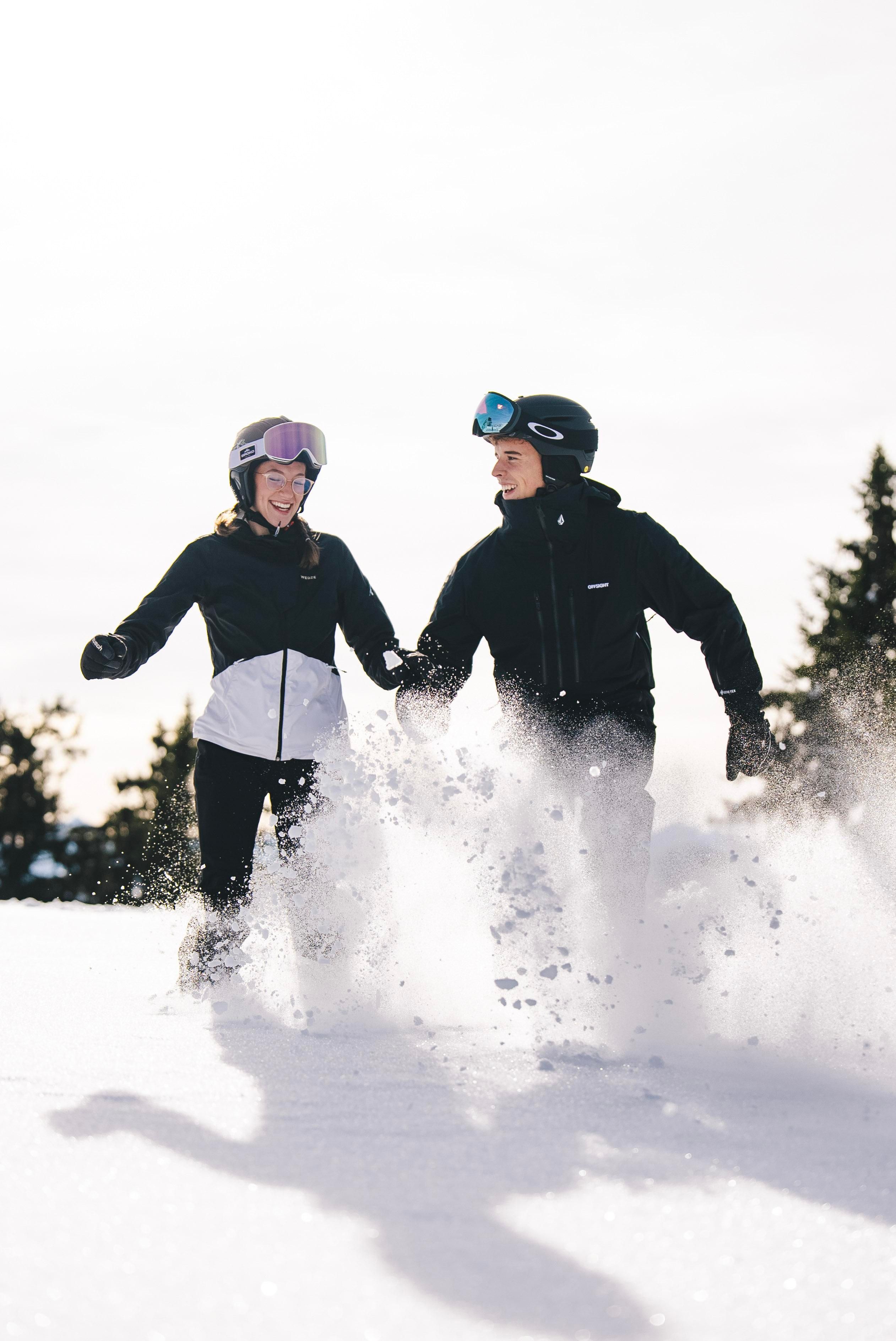 A couple in the snow is having fun and playing together. Both are wearing ski gear and laughing as they walk through the fresh snow.