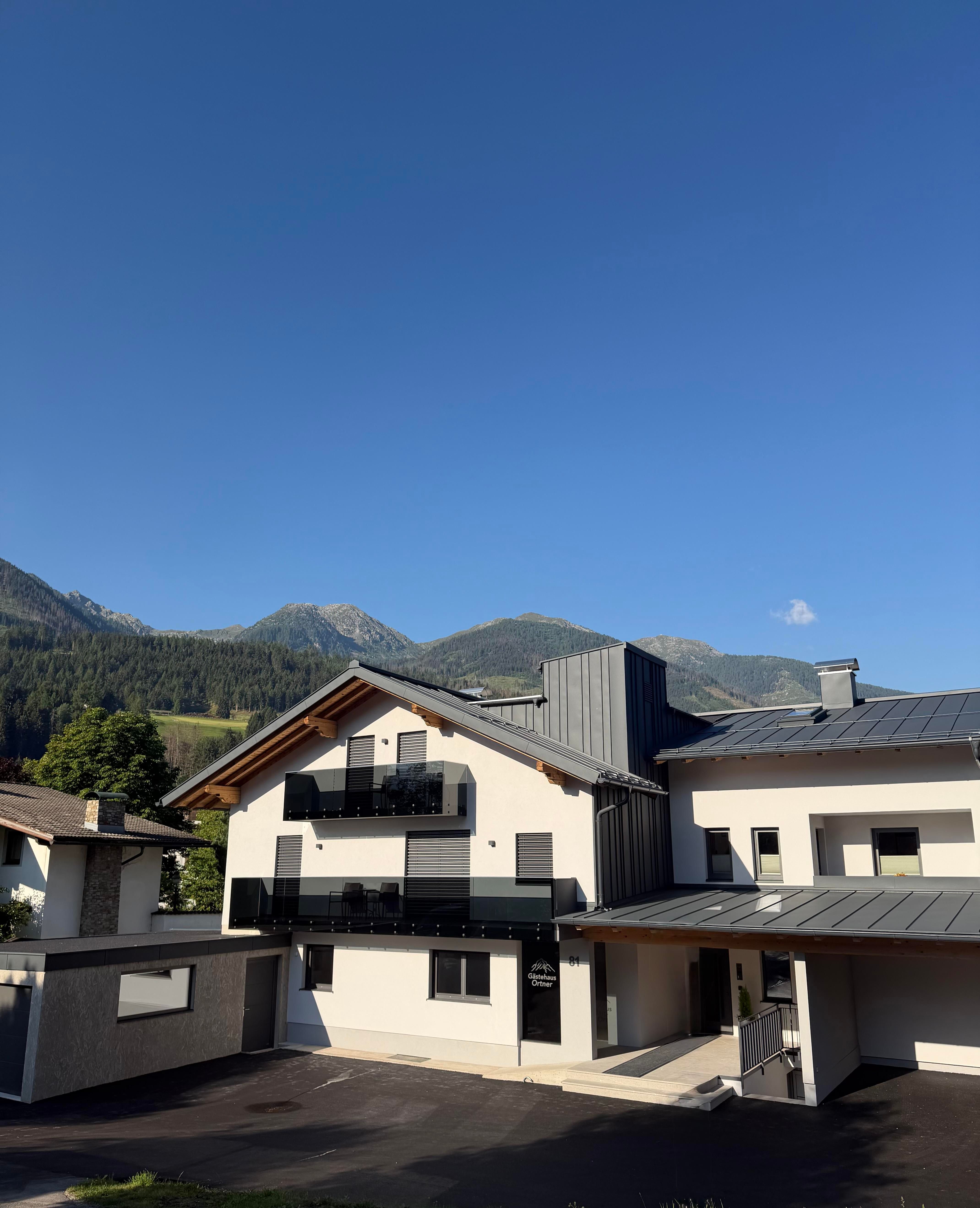 A modern building in a mountain landscape. The sky is clear and blue, while the mountains are visible in the background. Guesthouse Ortner from the front with the entrance area and parking spaces directly in front of the house.