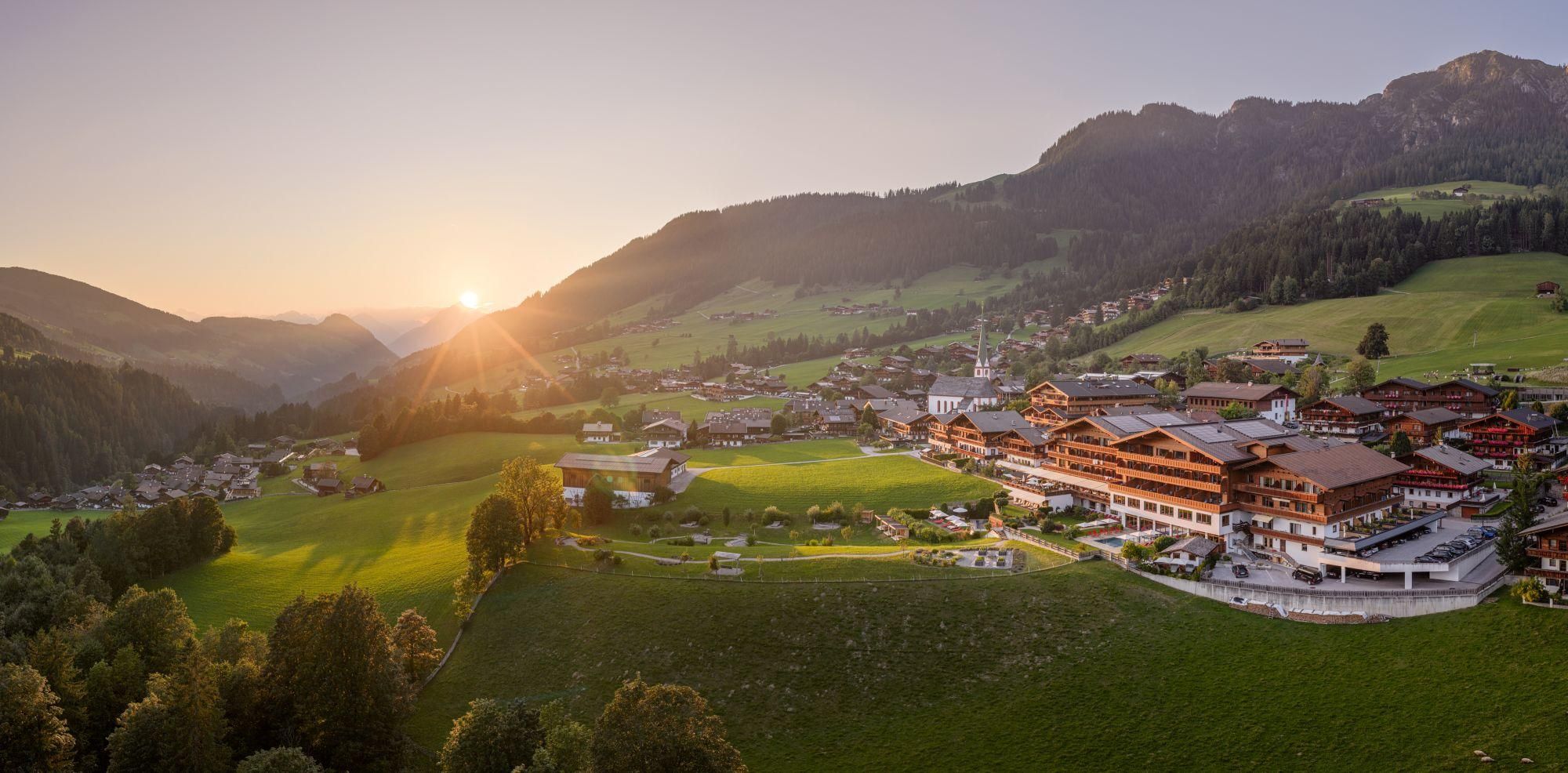 A picturesque mountain landscape with gentle hills and a small village. The sun rises above the mountains, bathing the scene in warm light.