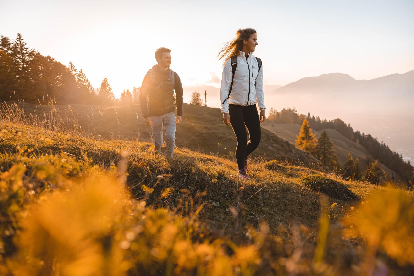 Two hikers enjoy nature at sunset. In the background, mountains and forests can be seen.