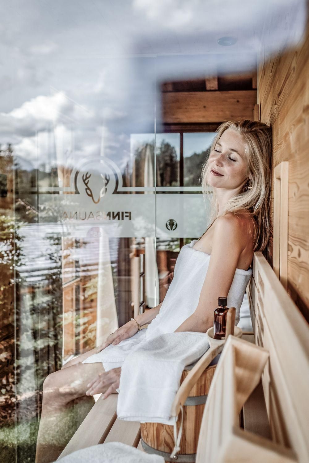 A relaxed woman is sitting in a sauna and enjoying the tranquility. Through the window, there is a beautiful view of nature.