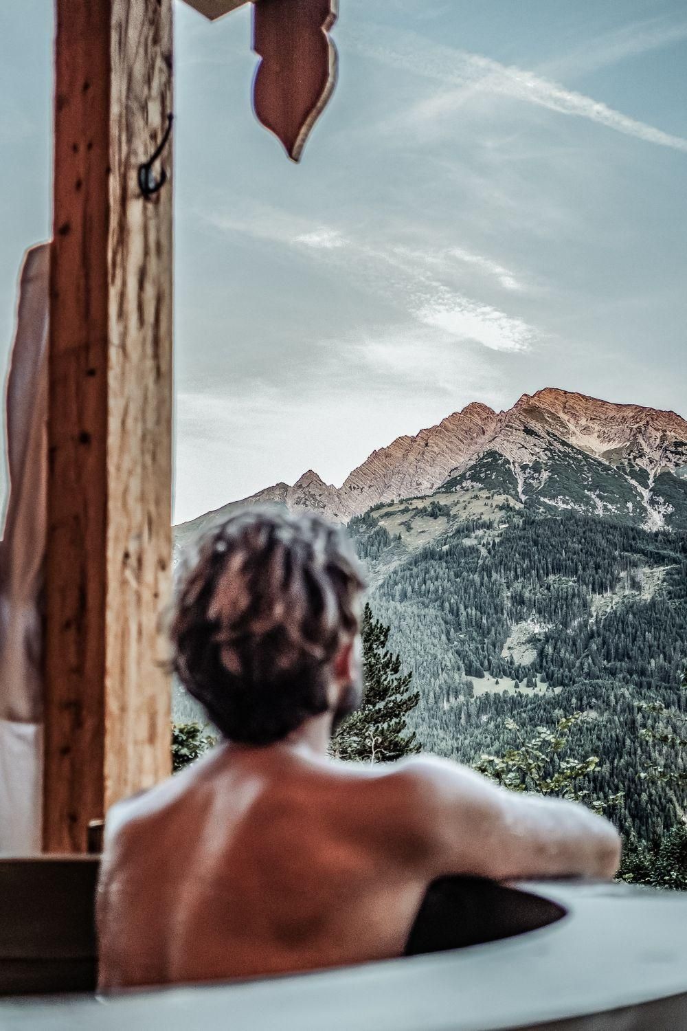 A person is sitting relaxed in a hot pot and looking at the majestic mountains. The sky is clear and nature appears peaceful.