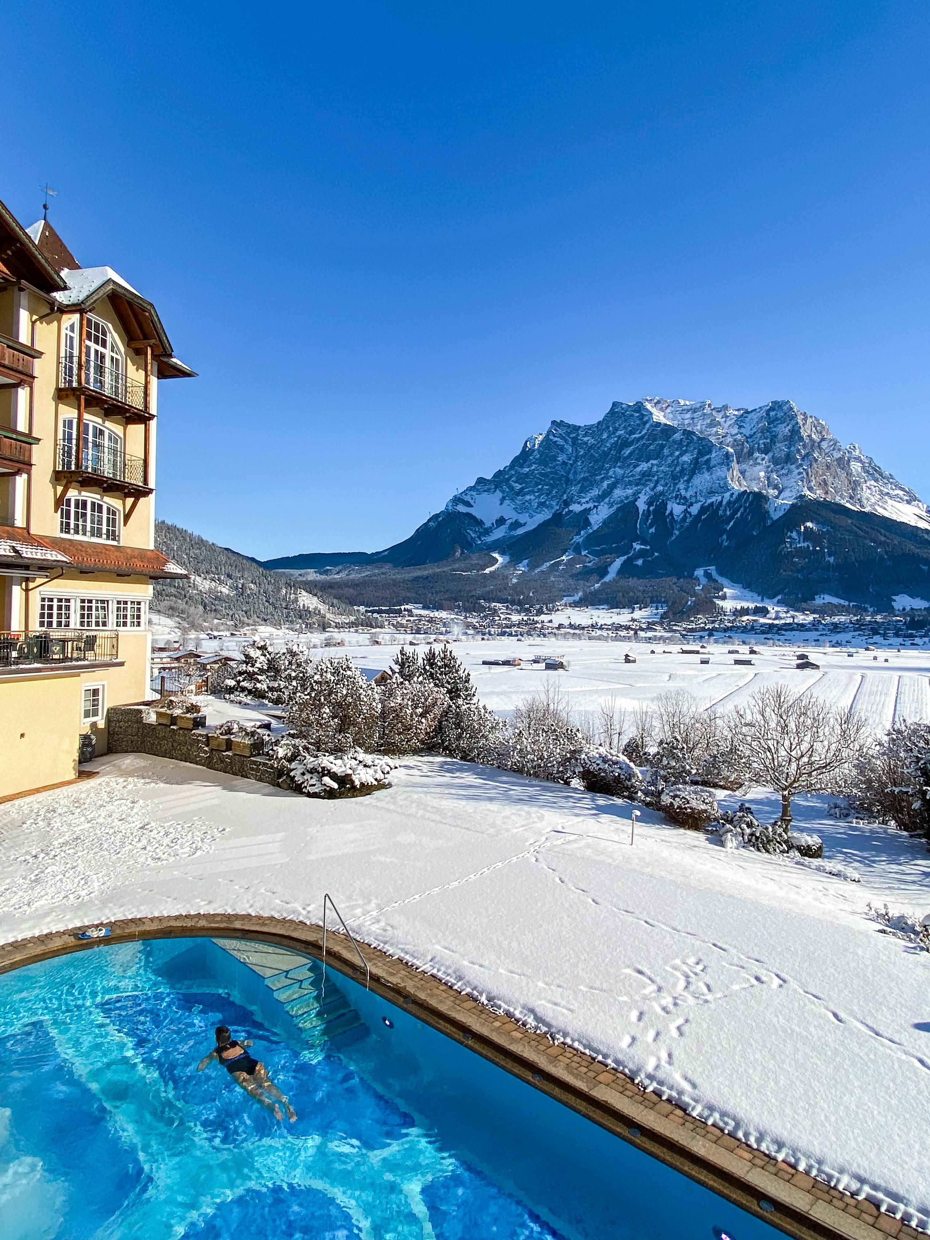 A beautiful pool with a person swimming, surrounded by snow-covered landscape. In the background, a majestic mountain rises under a clear blue sky.