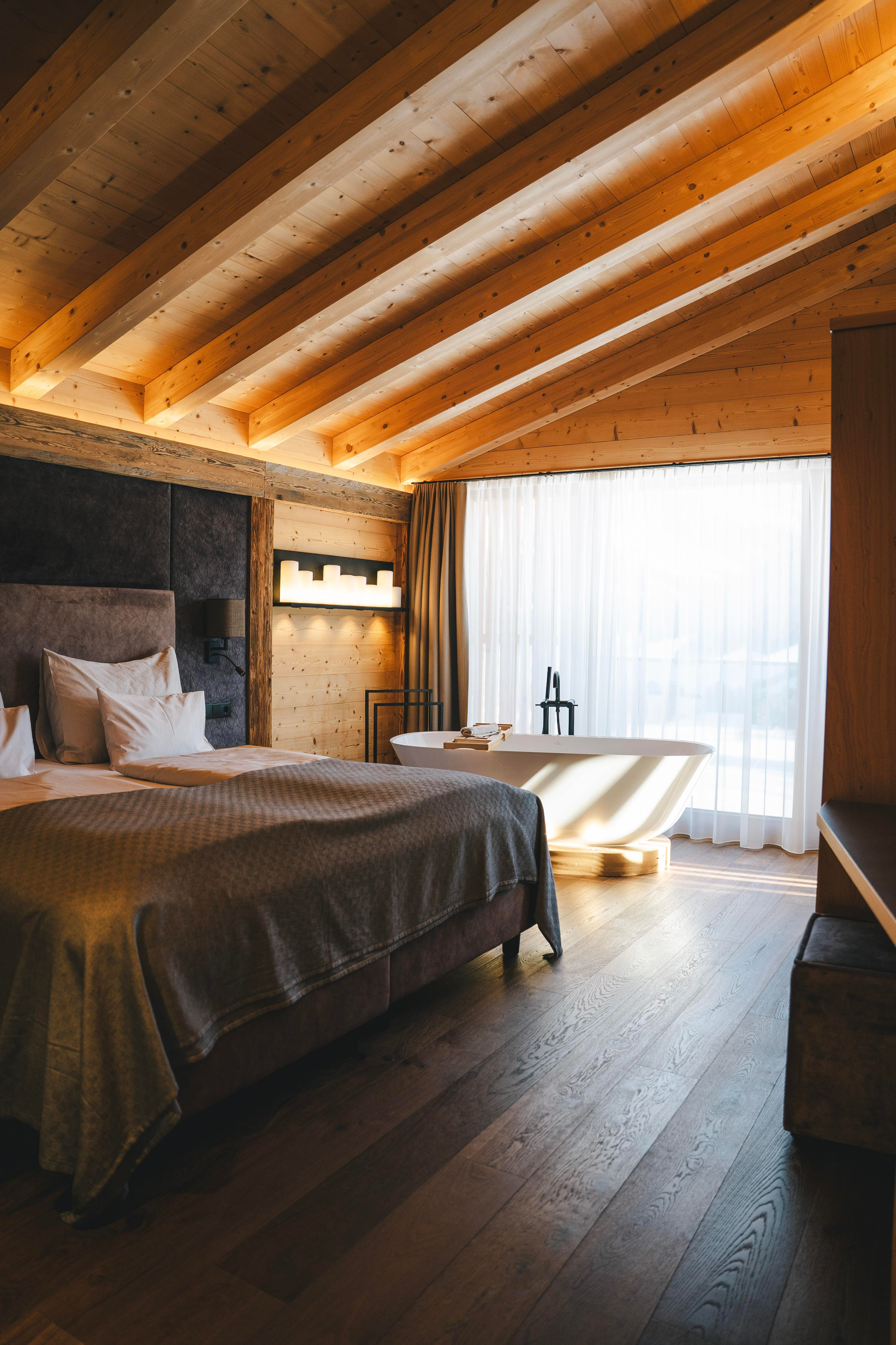 A modern bedroom with wooden ceilings and a large window. The room includes a bed, a bathtub, and a stylish decor.