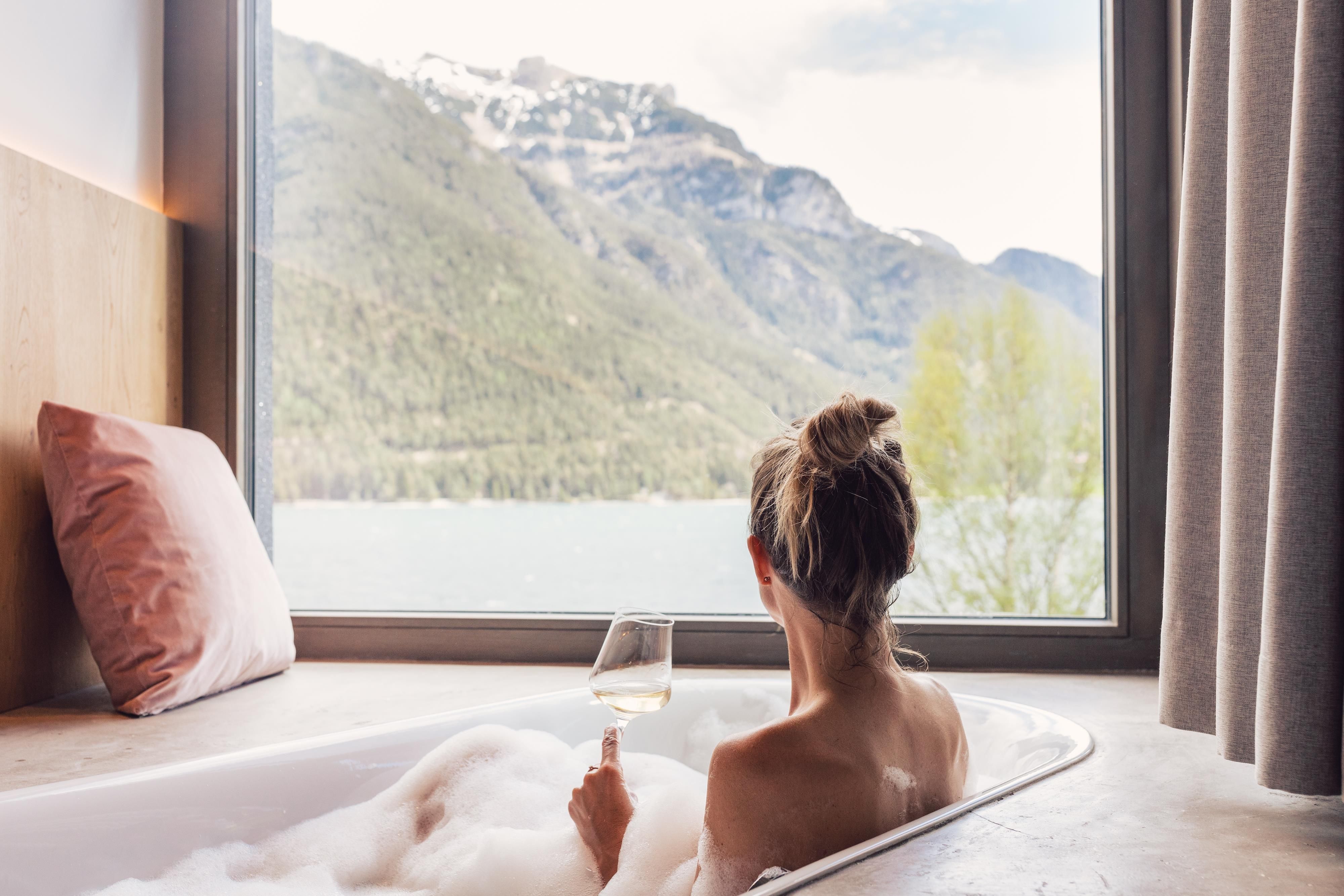 A person is sitting in a bathtub, enjoying a view of the mountains and a lake. They are holding a glass in their hand and surrounded by泡沫 and cushions.