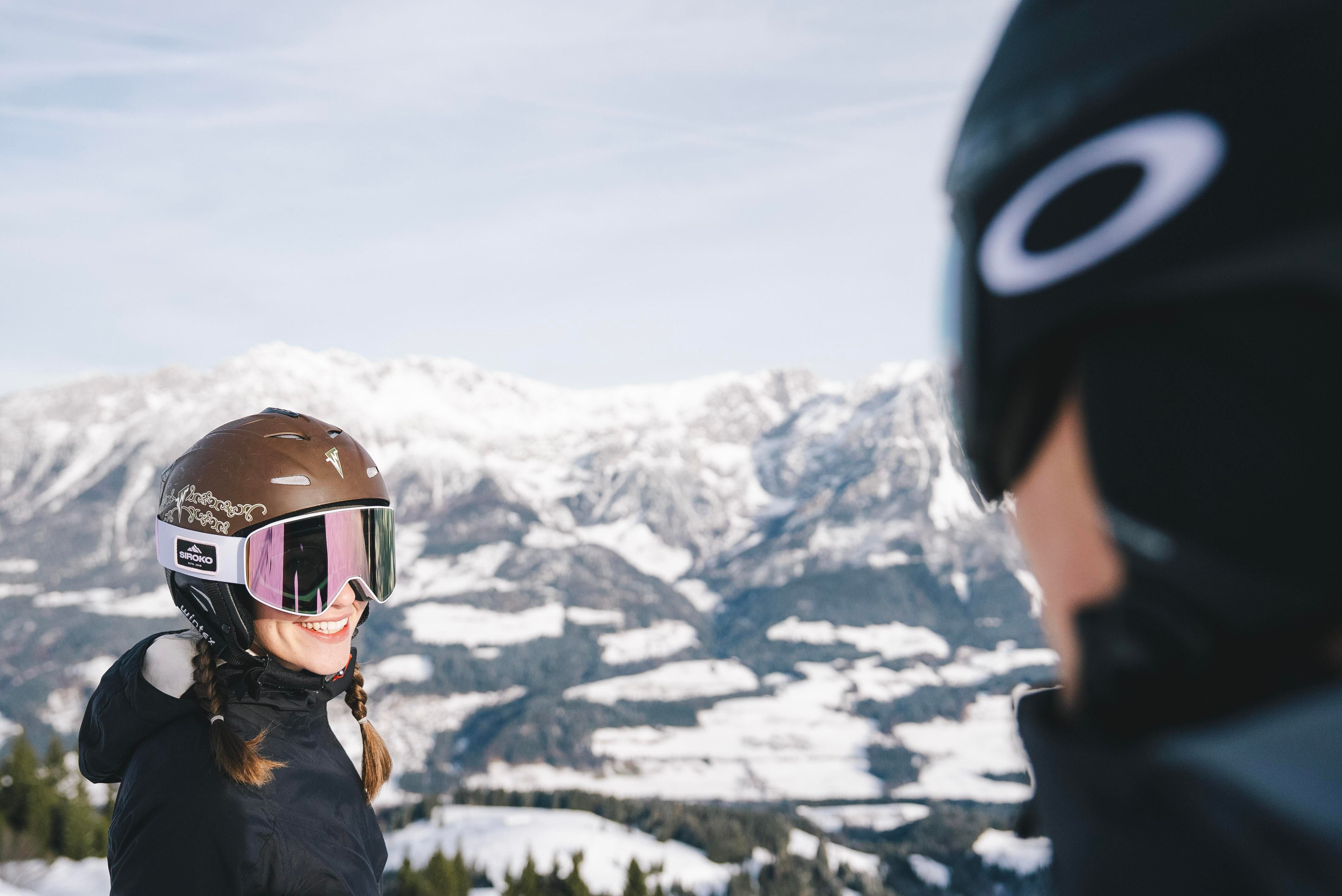 Two skiers are smiling at each other on a snow-covered mountain. In the background, majestic mountains and a clear sky can be seen.