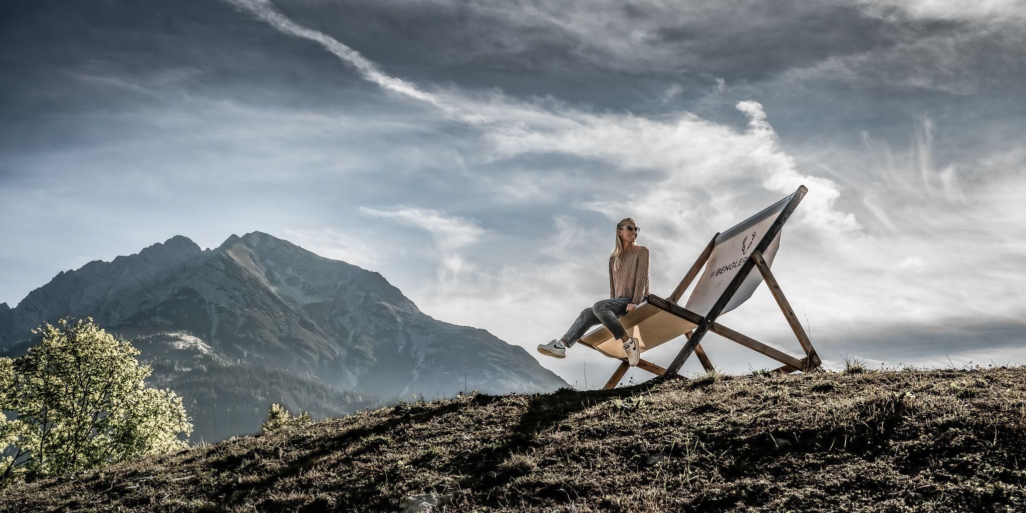 A woman is sitting relaxed on a lounge chair in nature. In the background, there are mountains and a cloudy sky.