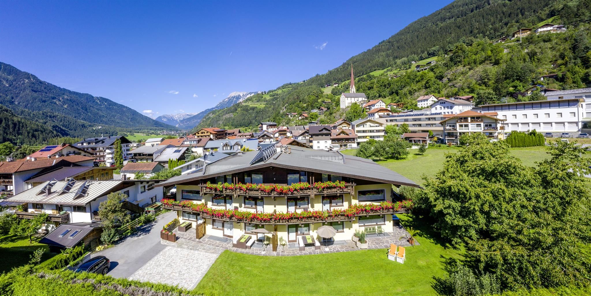 A picturesque mountain landscape with a village in the valley. In the foreground, there is a building with balconies and colorful flowers.