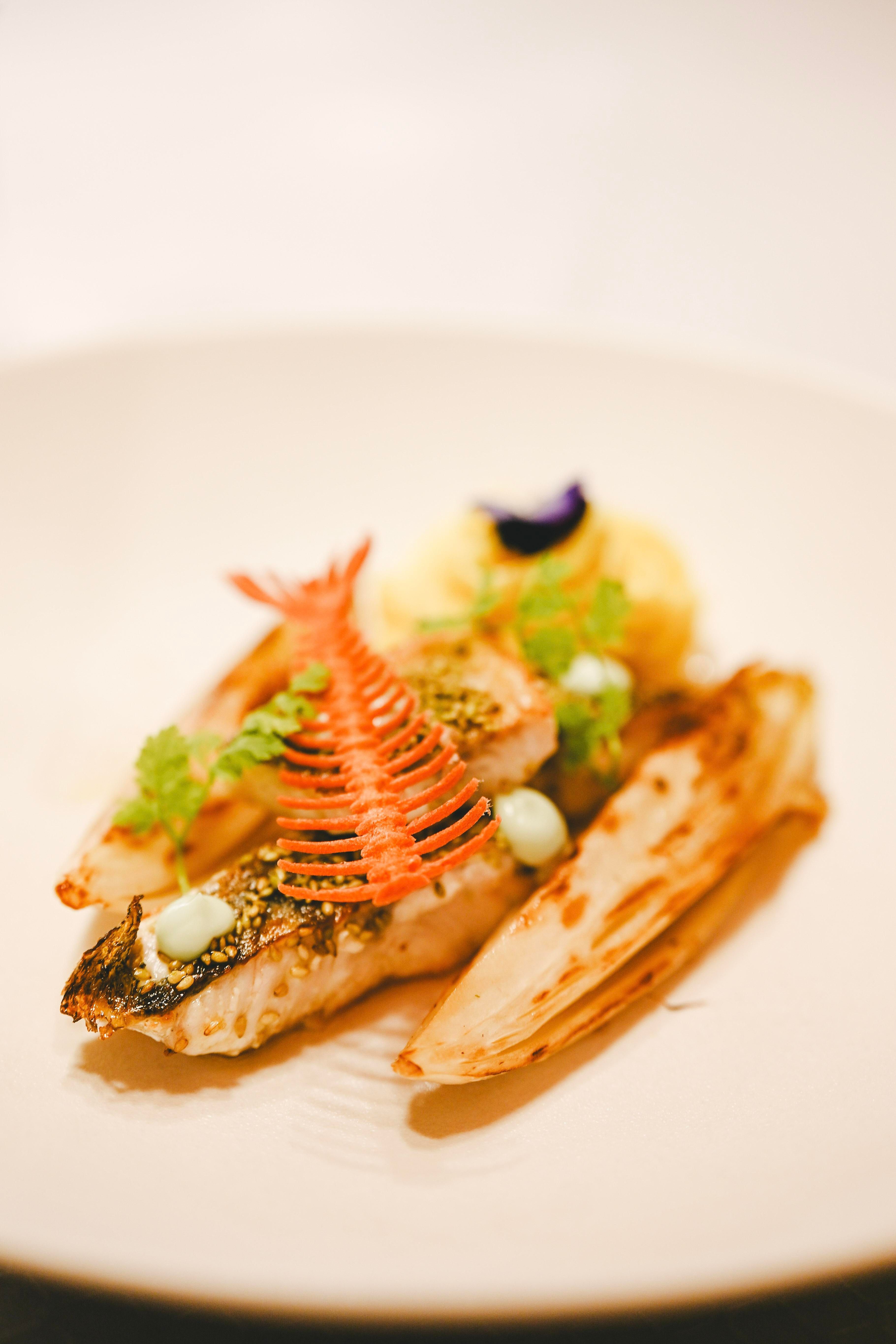 A artfully arranged fish on a white plate, garnished with fresh herbs and colorful decorations. Next to it, grilled vegetable sides can be seen.