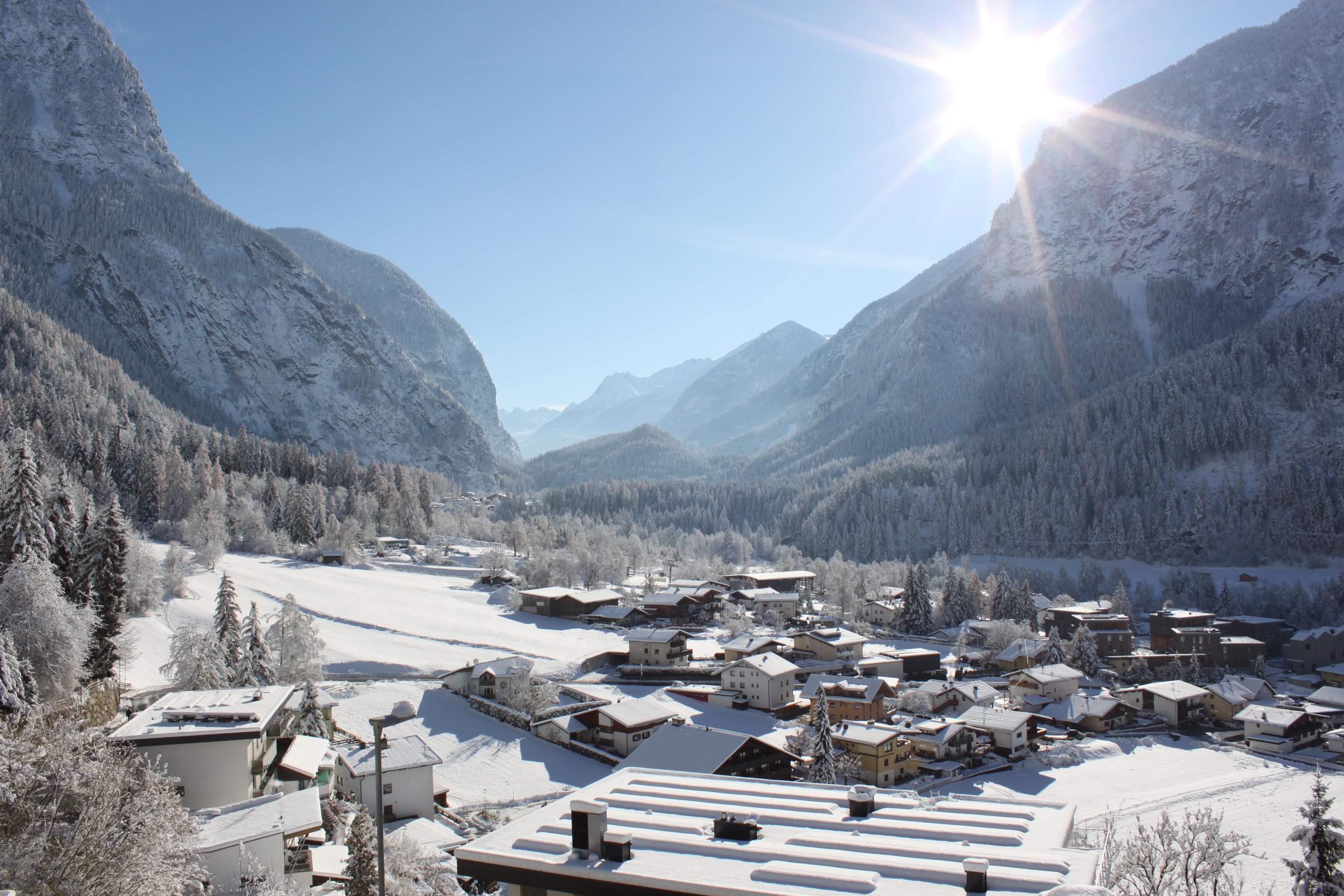A snowy landscape with a small village and high mountains in the background. The sun shines bright in the sky and illuminates the wintry scene.