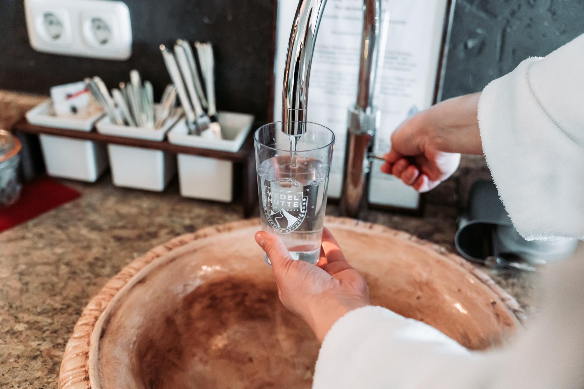 A hand is filling a glass with water from a faucet. In the background, utensils and a sink are visible.
