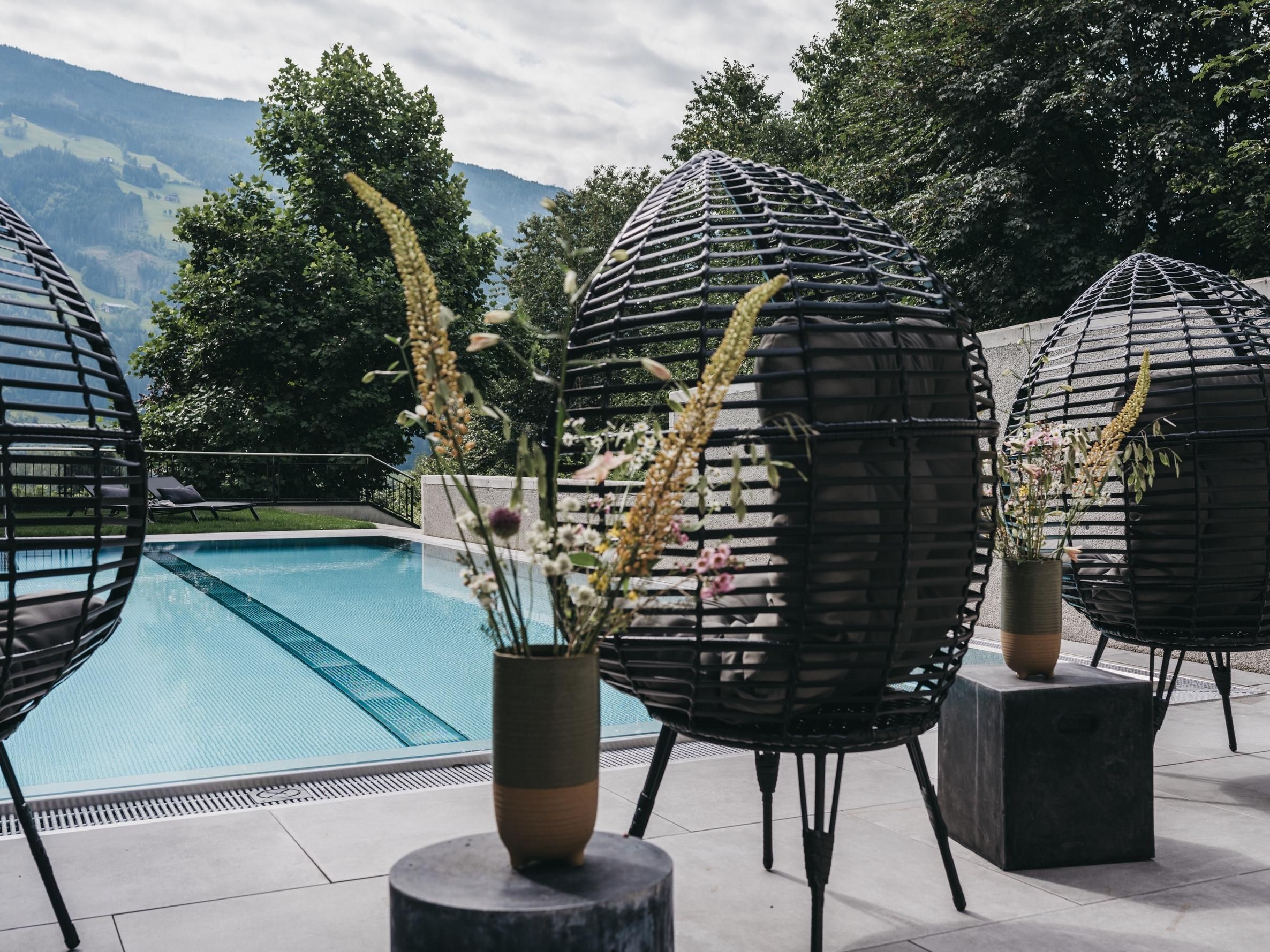 Modern seating in the shape of eggs is located at the edge of a pool. In the background, there are trees and a picturesque mountain landscape.