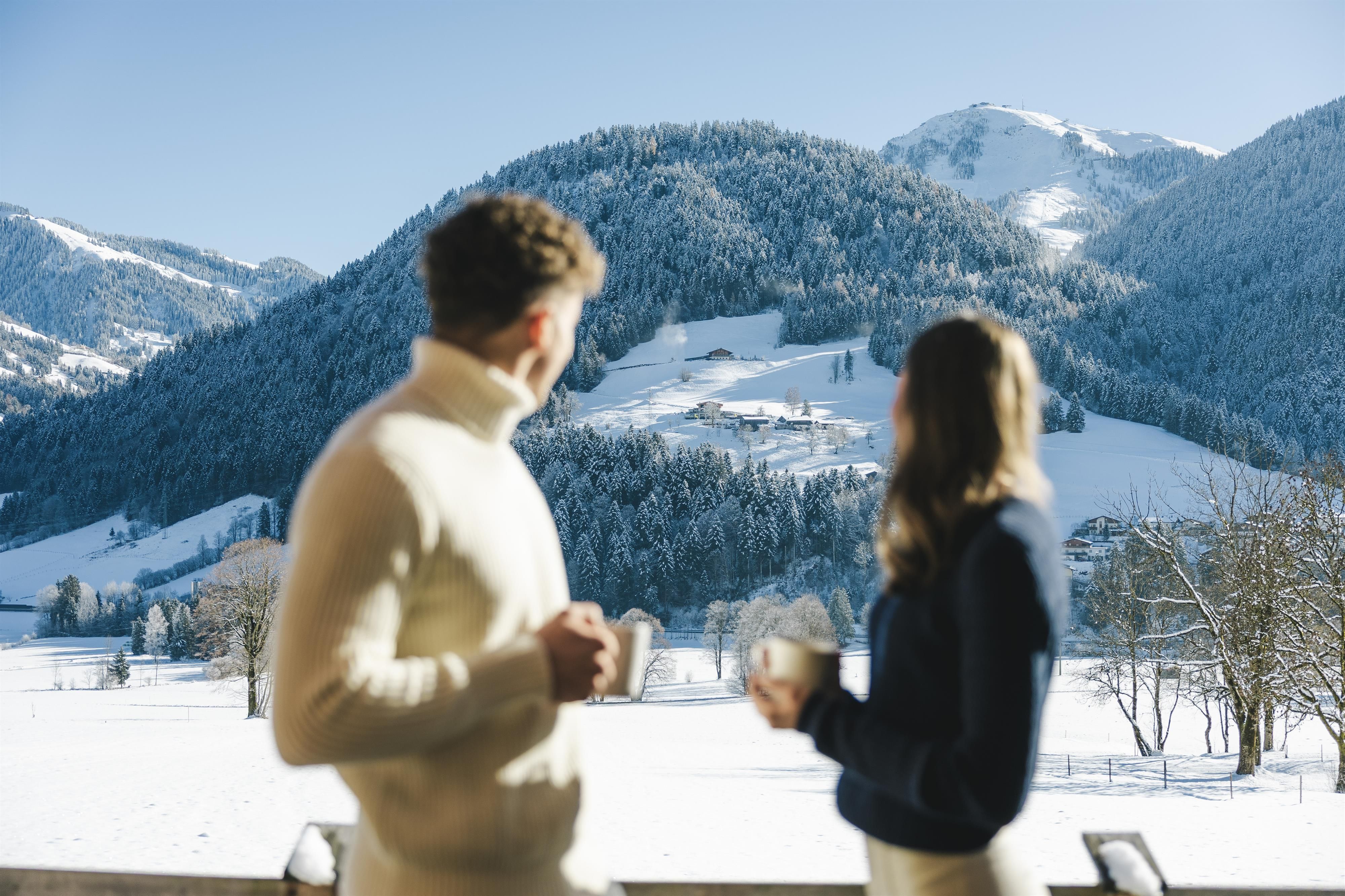A couple stands on a balcony enjoying hot drinks. In the background, a snowy mountain landscape stretches out under a clear blue sky.