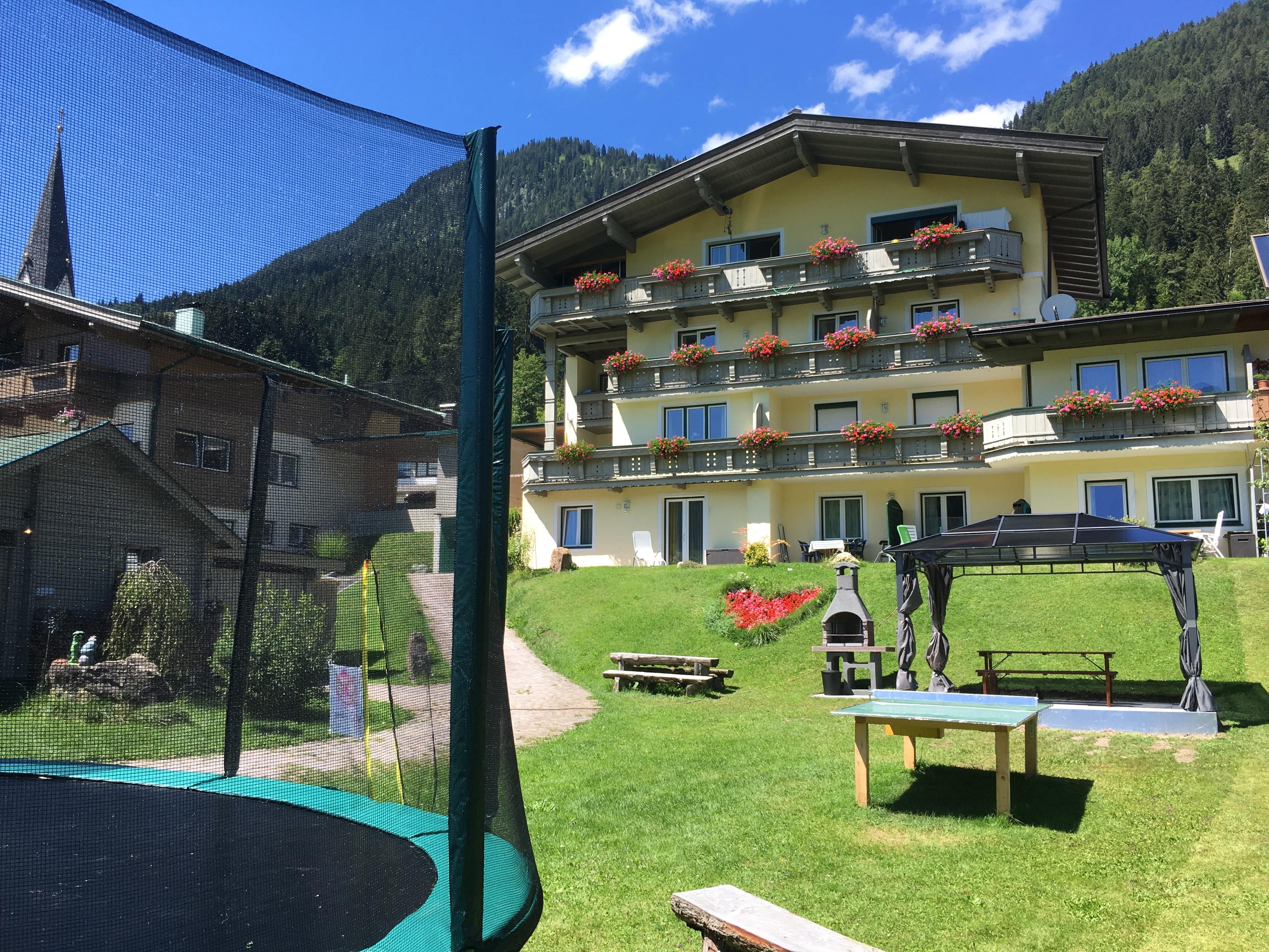 A cozy hotel building with balcony flowers in a picturesque mountain landscape. In the foreground, there is a trampoline and several outdoor seating areas.