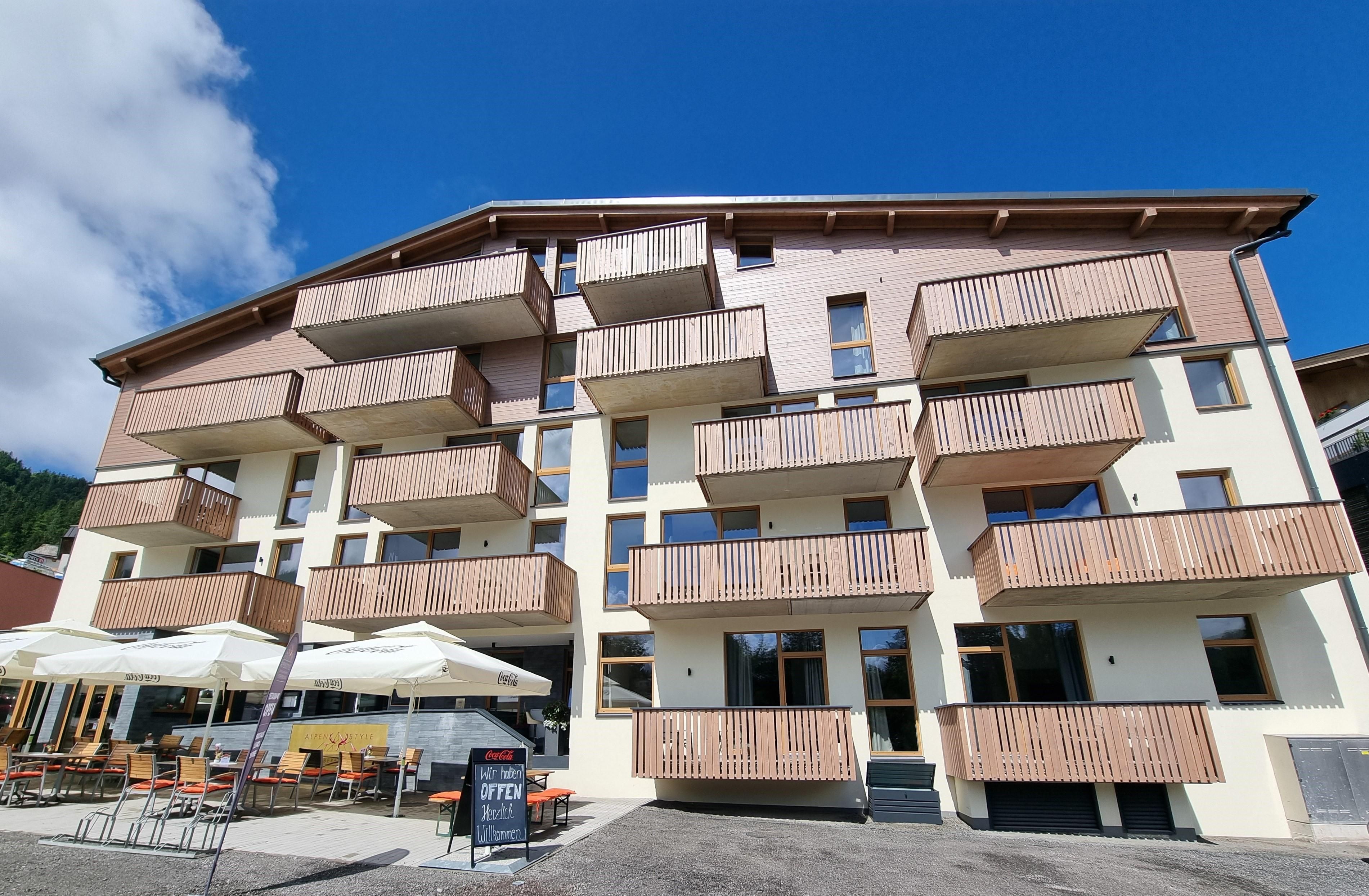 A modern building with balconies in bright colors. In front of the house, there are terraces with sun umbrellas.