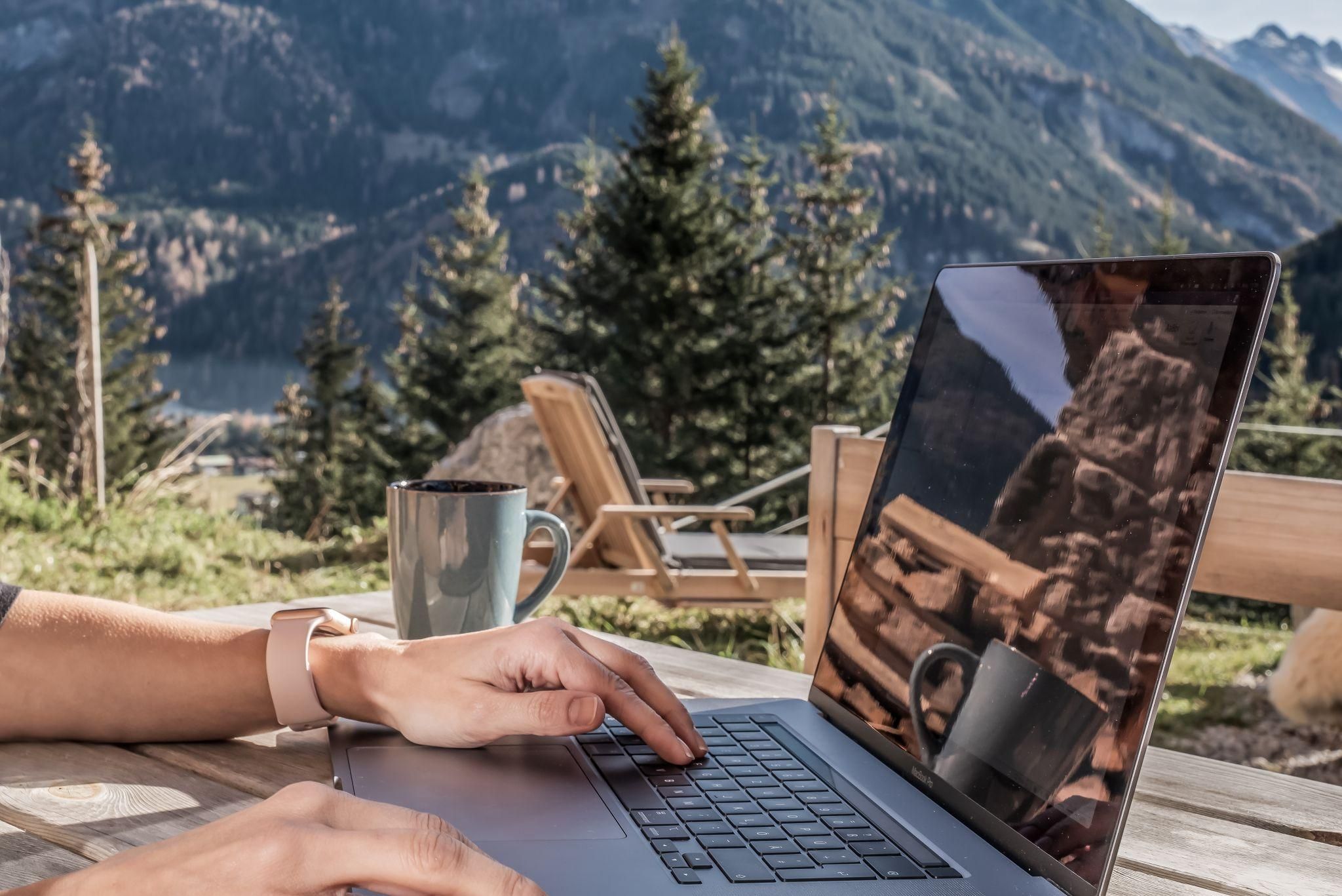 A person is working on a laptop outdoors, surrounded by beautiful mountains and trees. Next to the laptop is a cup of coffee.