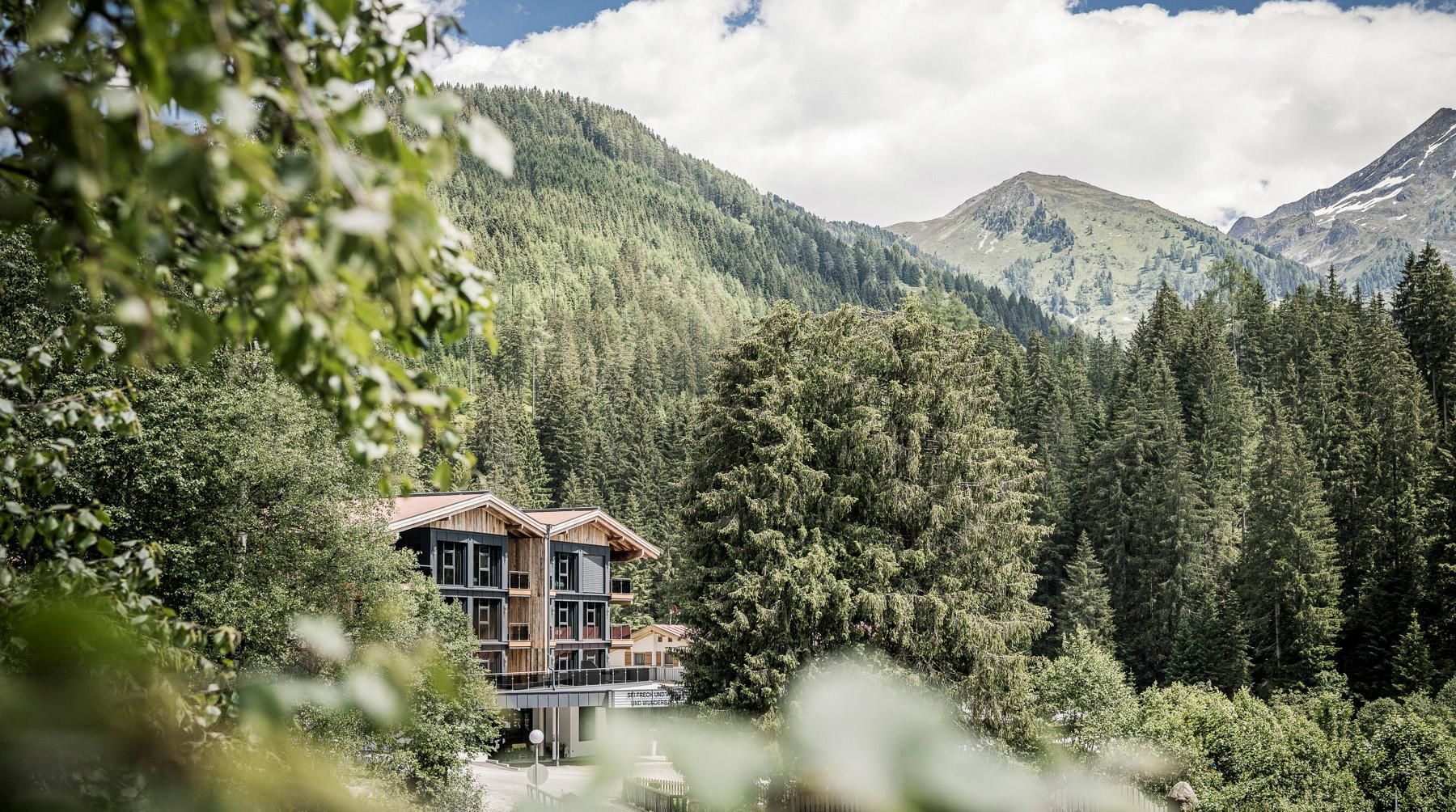 A modern house amidst tall trees and mountains. The sky is partly cloudy and the landscape appears calm and idyllic.