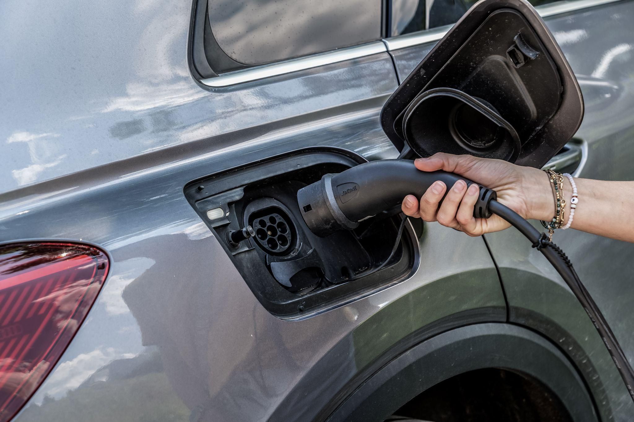 A hand plugging a charging cable into the charging port of a gray electric vehicle. The background shows the design of the car body.