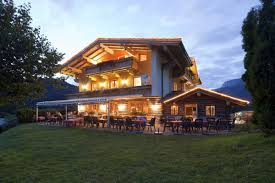 A cozy house in alpine style, illuminated at dusk. In the foreground, there is a terrace with outdoor seating.