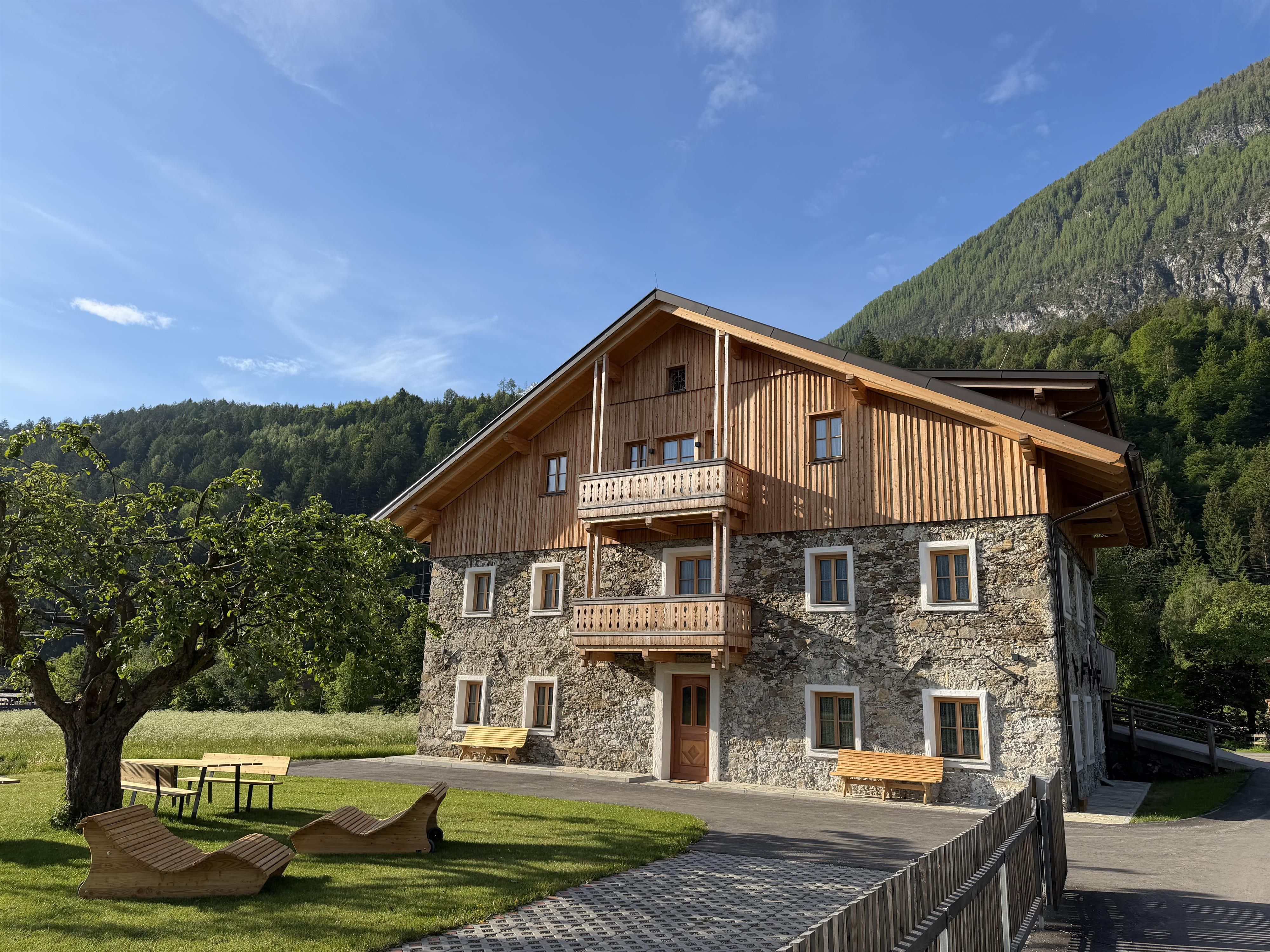Old farmhouse surrounded by nature and mountains. In the foreground you can see loungers on a lawn and an apple tree.