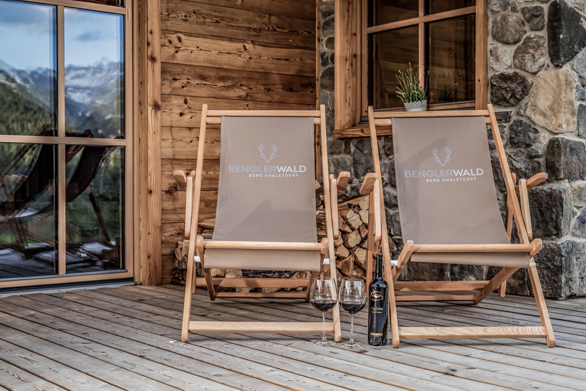 Two relaxing lounge chairs are placed on a wooden terrace in front of a chalet. Next to them are two wine glasses and a bottle of wine.