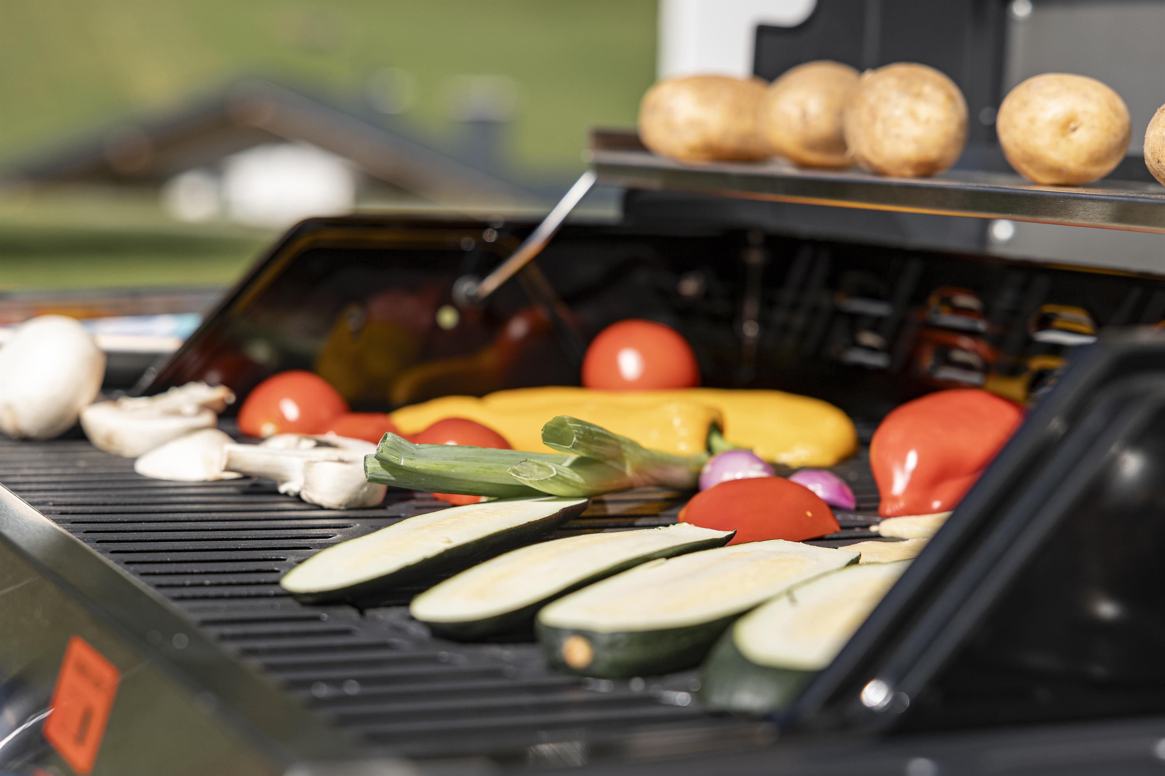 Colorful vegetables on a grill with fresh ingredients like zucchini, tomatoes, and bell peppers. In the background, potatoes are visible, being prepared for grilling.