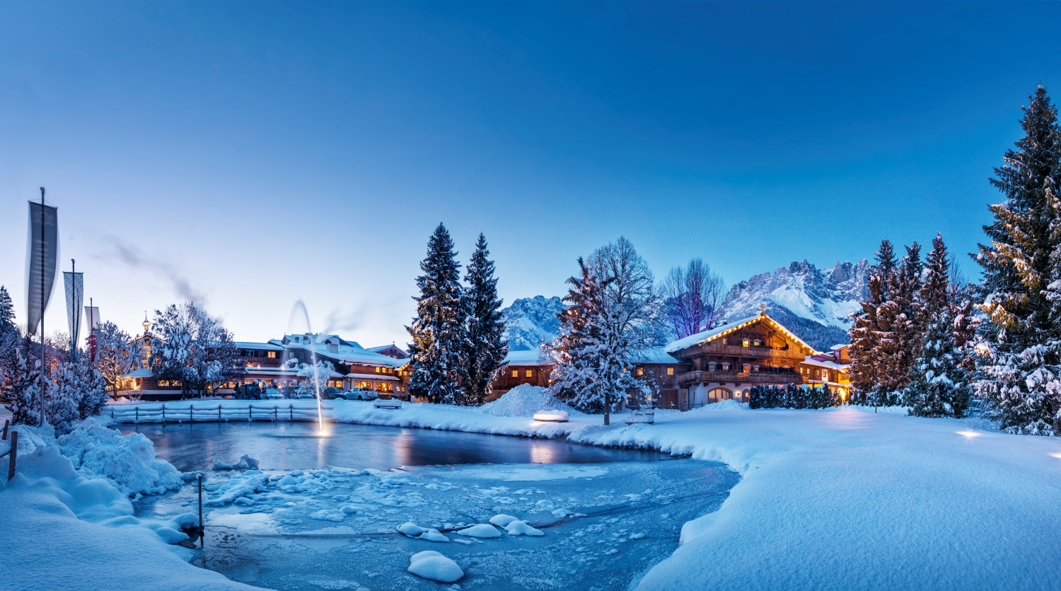 A picturesque winter landscape with snow-covered trees and a frozen pond. In the background, cozy cabins and mountains can be seen under a clear blue sky.