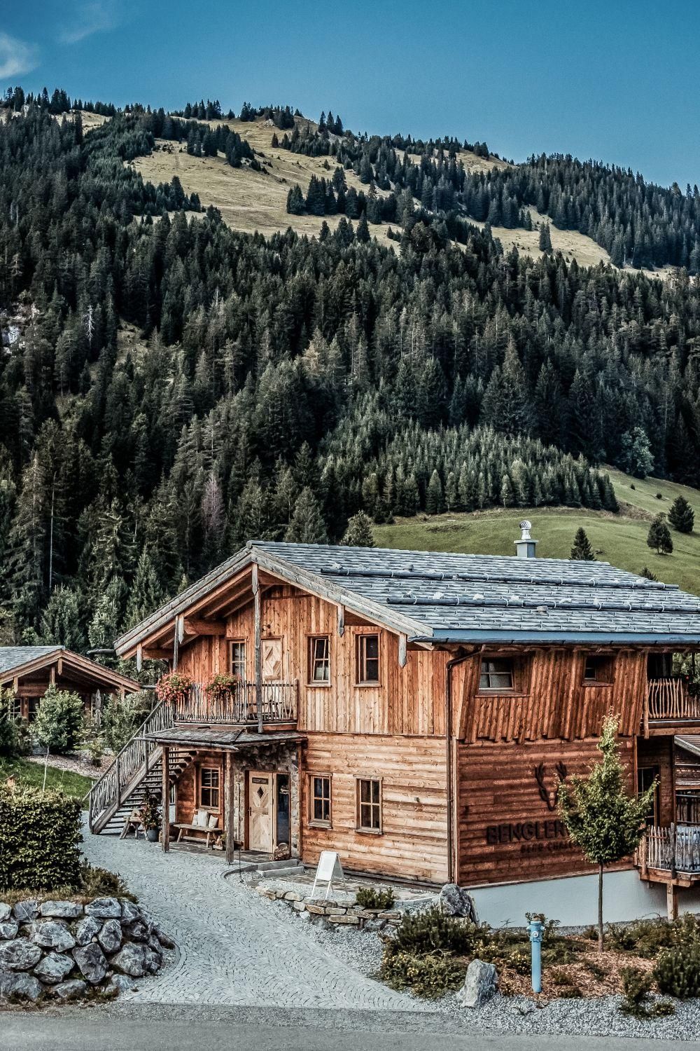 A cozy wooden house surrounded by green forests and mountains. The sky is clear and sunny.