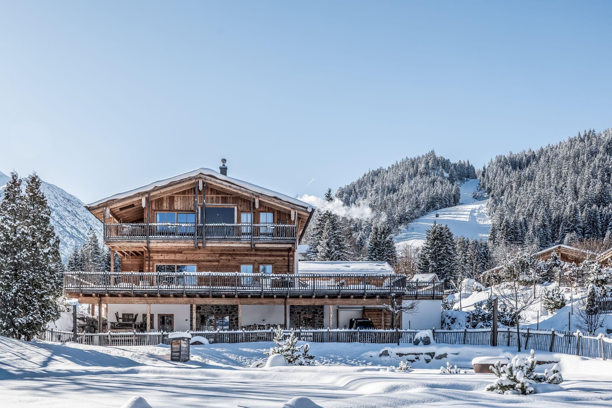 A charming chalet in winter covered with snow. The surrounding mountains are adorned with fir trees and the sky is clear.