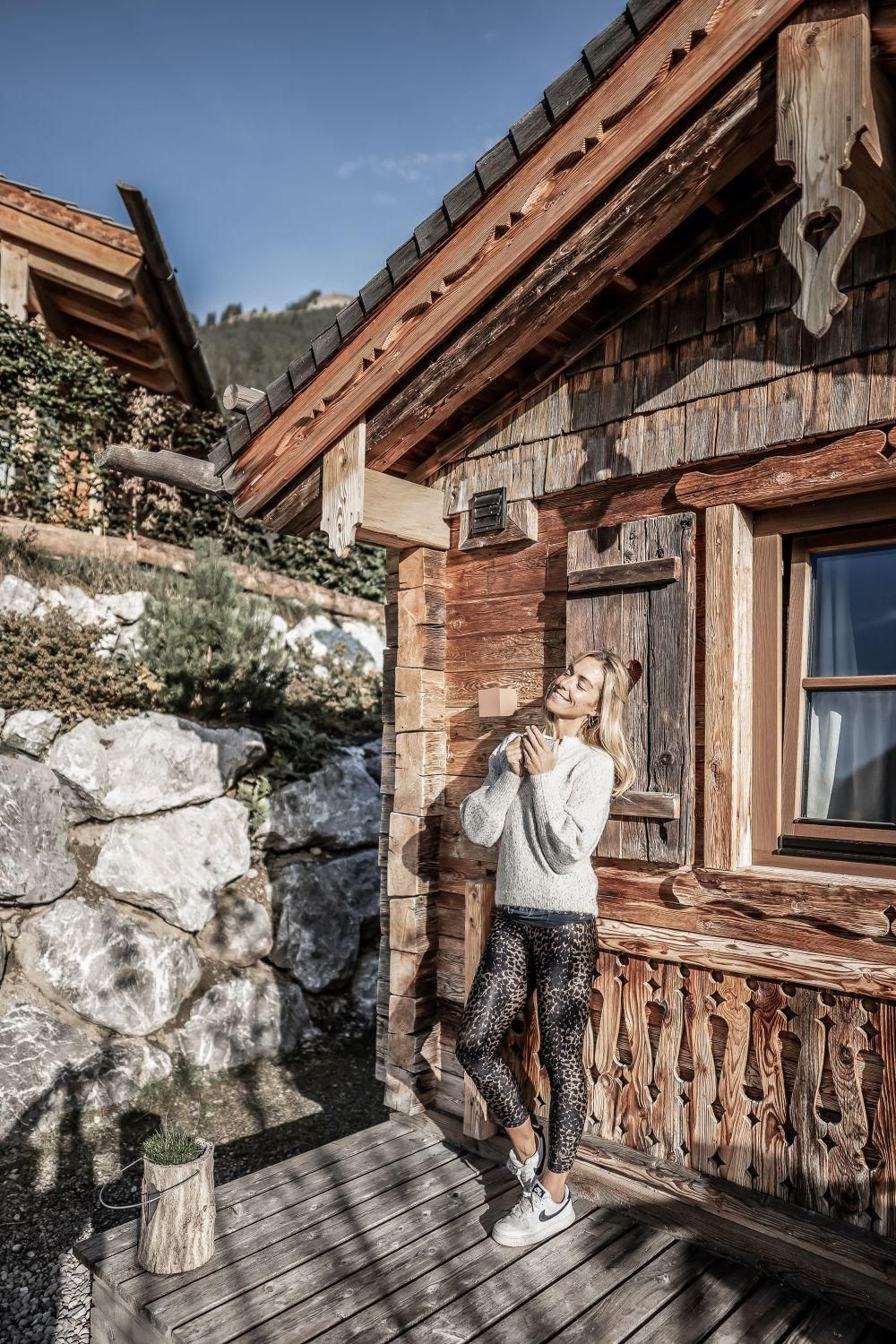 A young woman is standing relaxed in front of a chalet. In the background, rocks and green landscape can be seen.