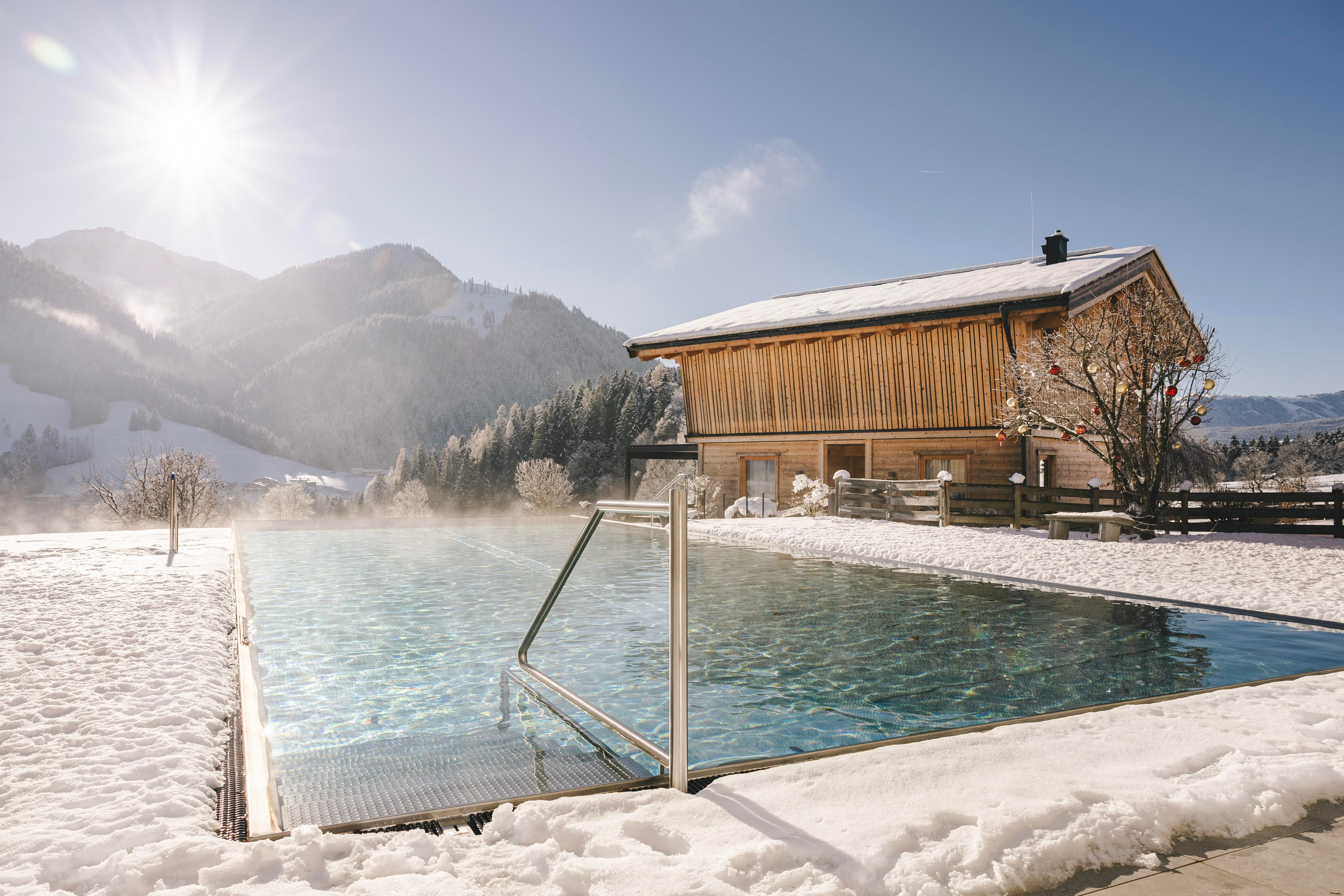 A tranquil outdoor pool in a snow-covered landscape. In the background, a wooden house and surrounding mountains can be seen, while the sun shines.