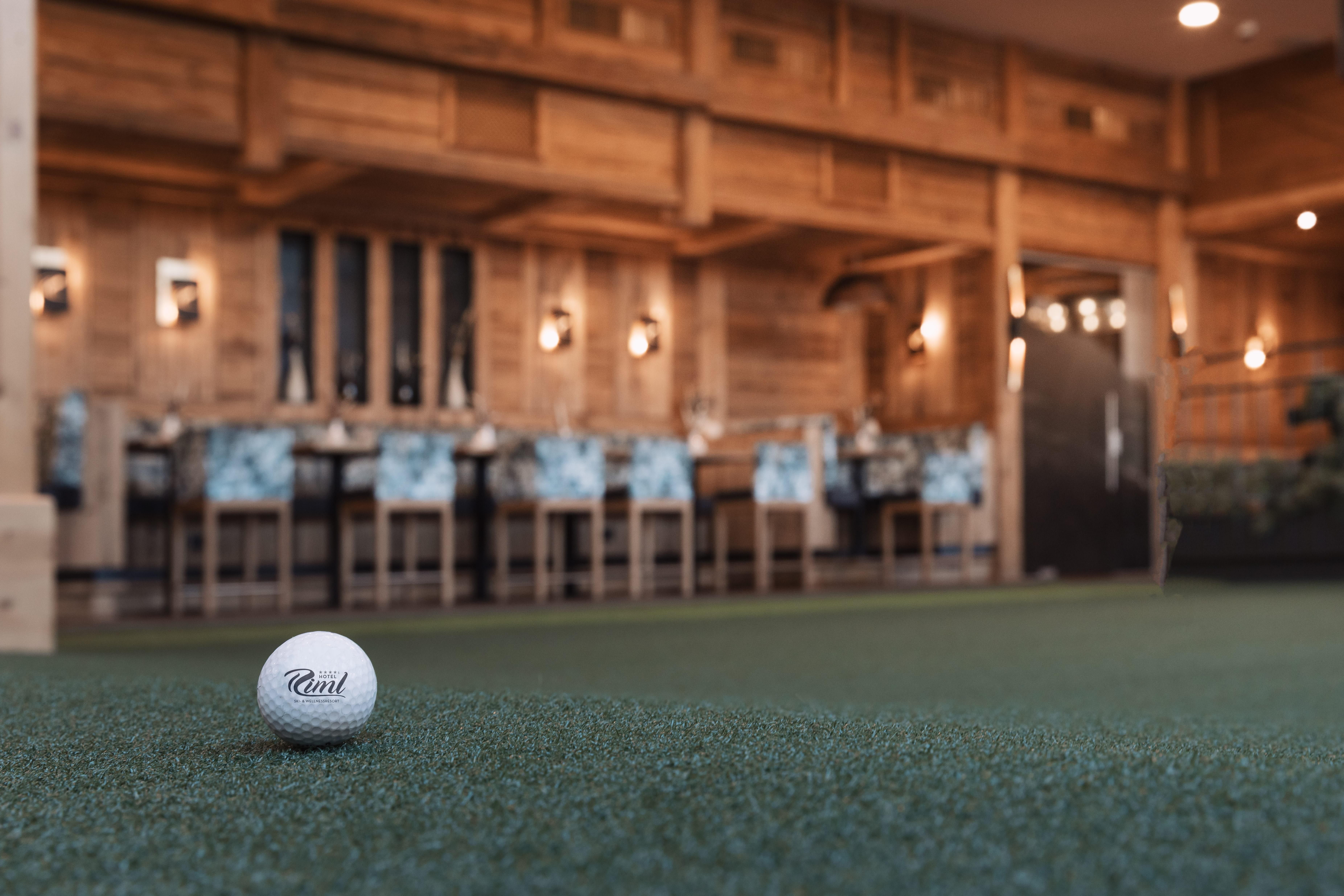 A golf ball lies on a green patch in the foreground. In the background, a rustic decor and tables can be seen in a cozy room.