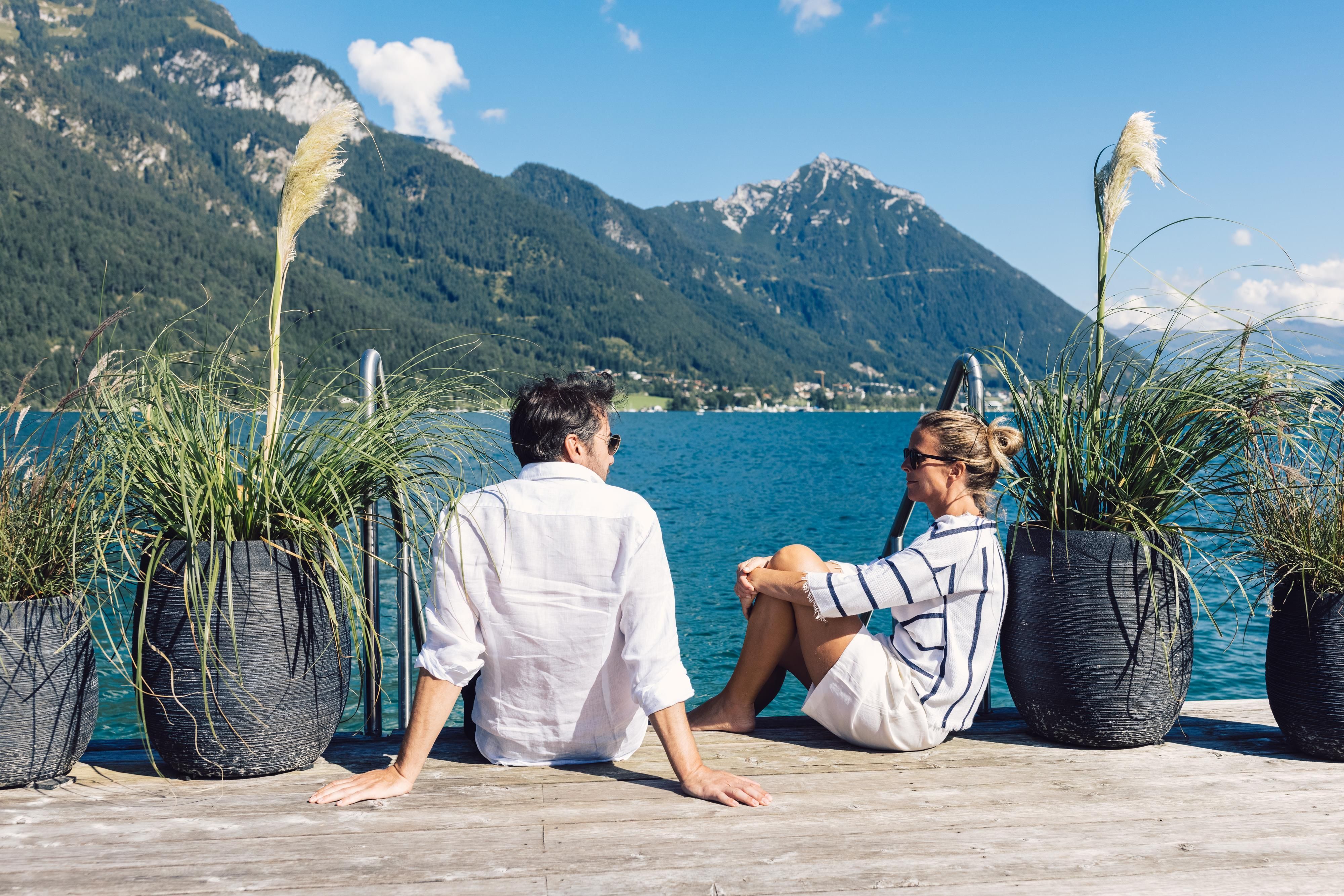 A couple sits by the shore of a lake and enjoys the view of the mountains. Large potted plants surround them.