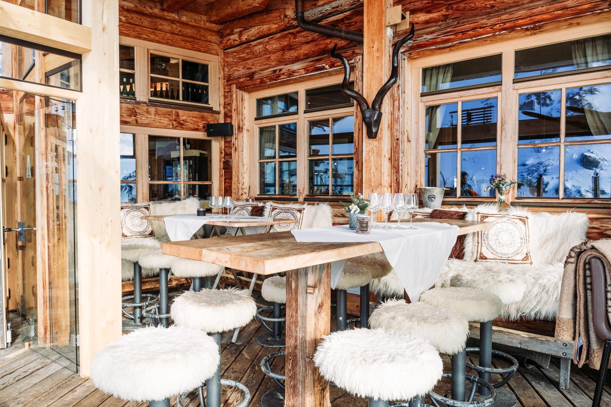 A cozy restaurant with wooden walls and large windows. The interior decor is rustic, with fur covers on the chairs and an inviting atmosphere.