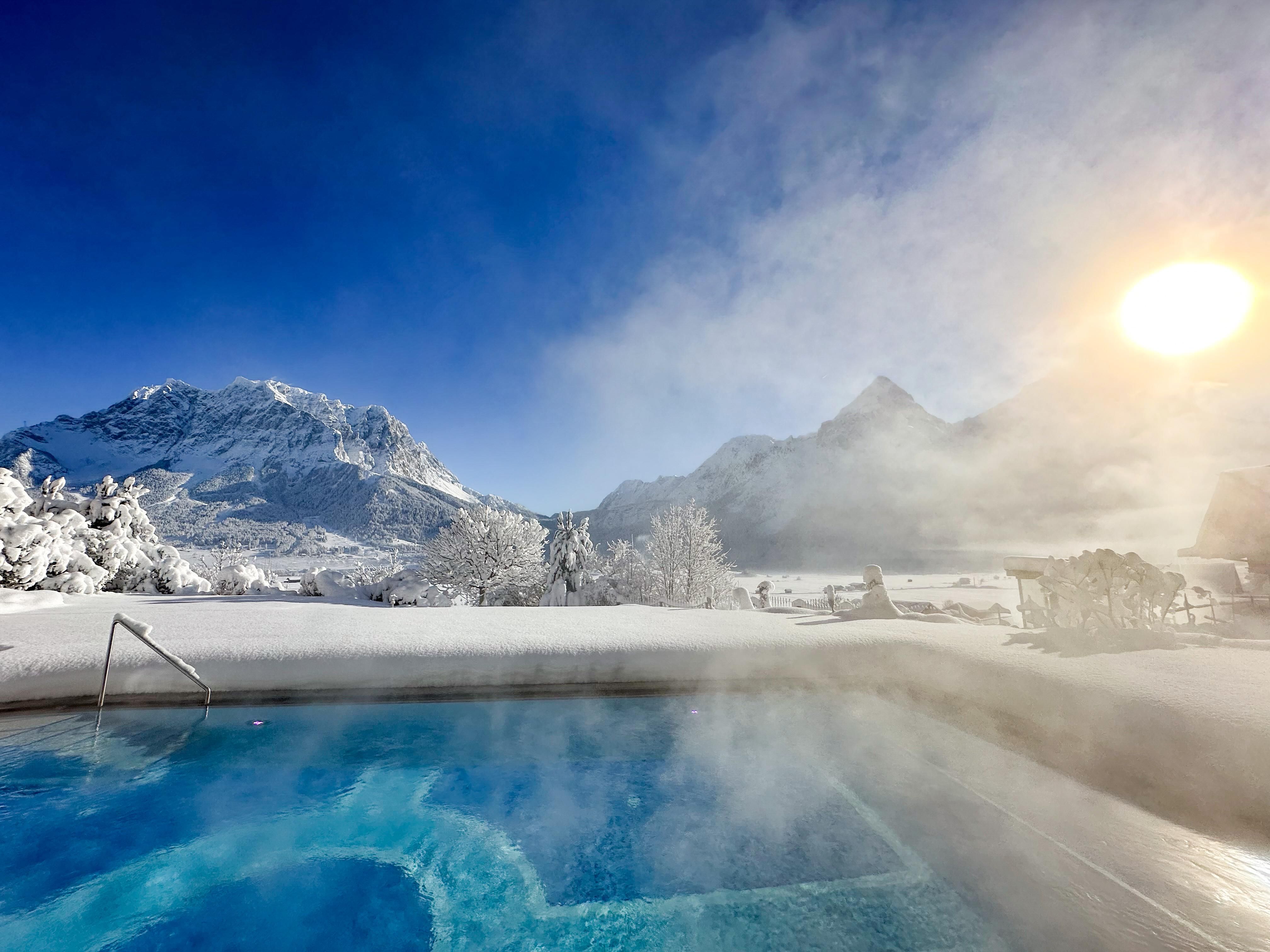 A beautiful outdoor pool surrounded by snow-covered mountains. The sun is shining and creating a steaming effect over the water.
