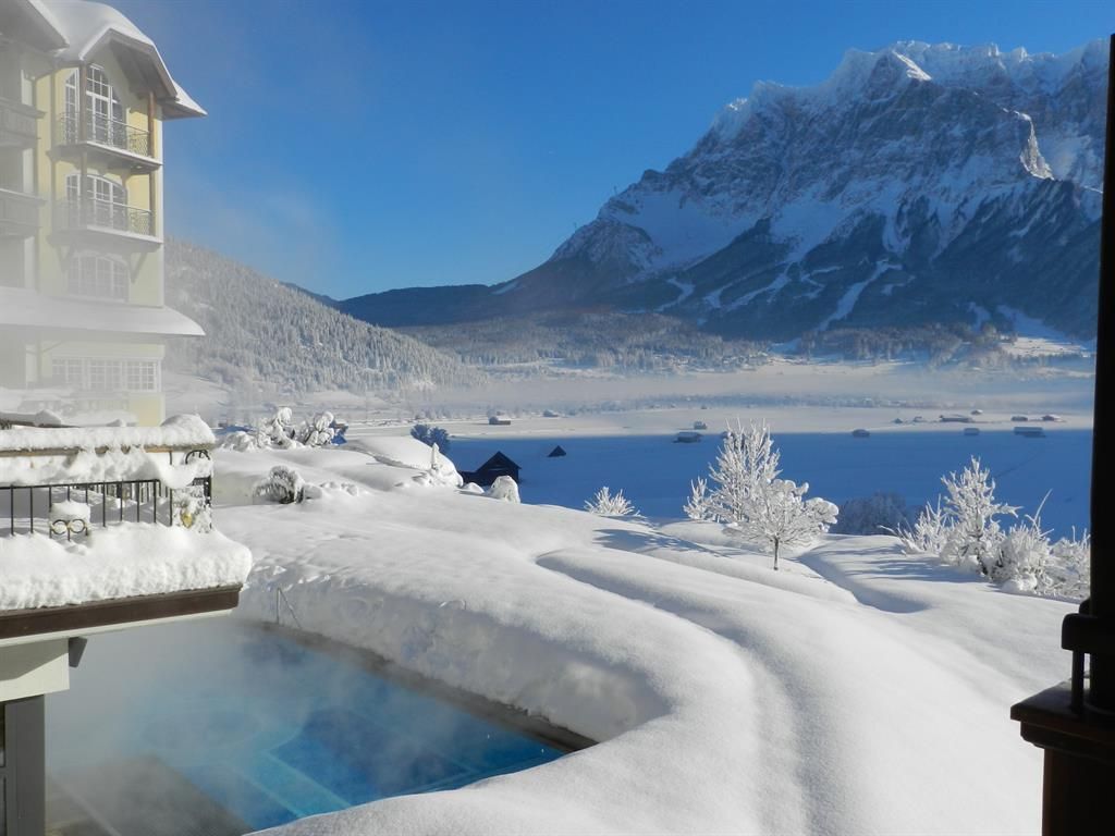 A picturesque winter landscape with snow-covered mountains and a clear sky. In the foreground, there is a warm outdoor pool surrounded by snow.