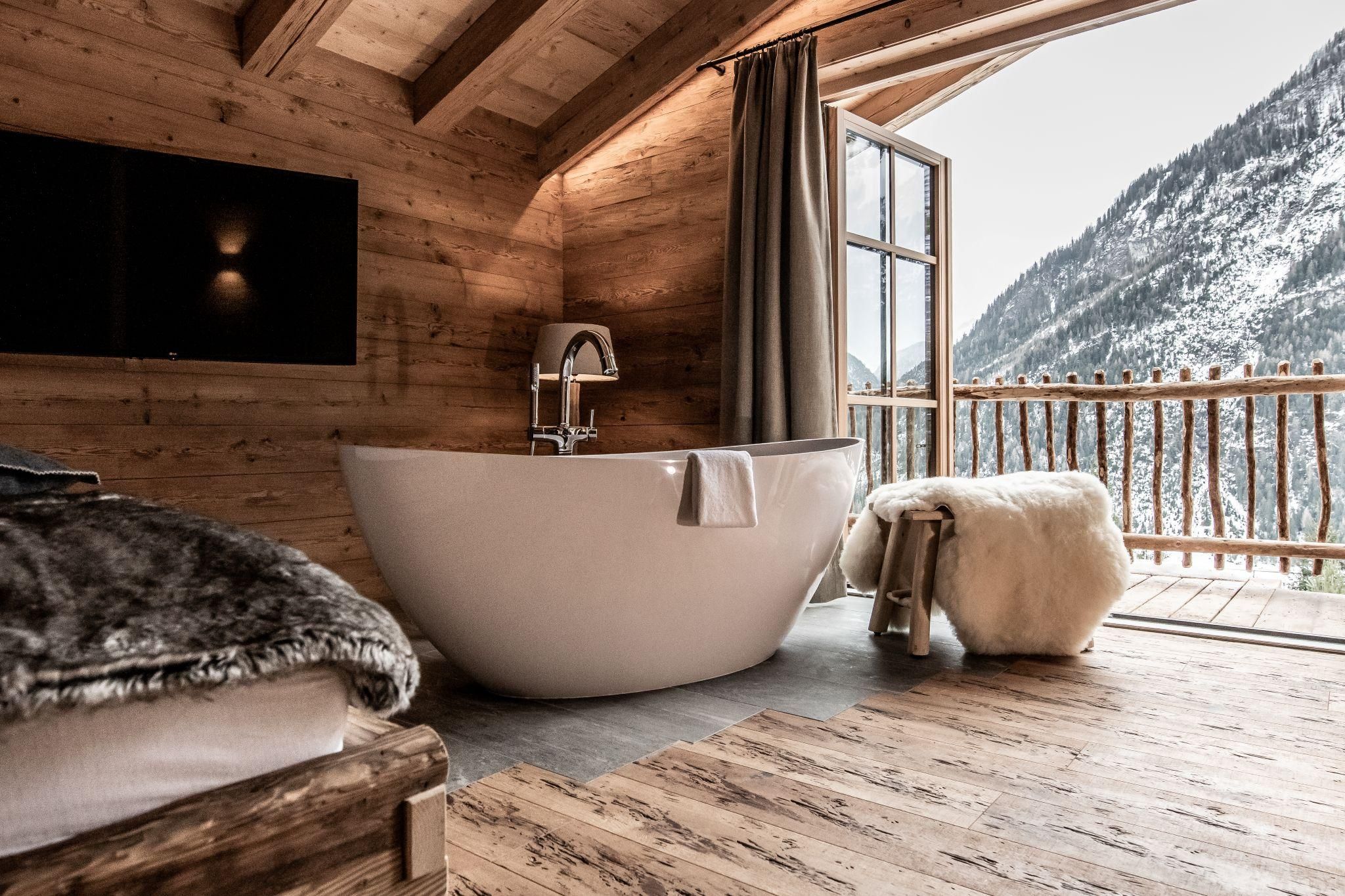 A modern bathroom in a chalet with a freestanding bathtub. The large window front offers a view of snow-covered mountains.