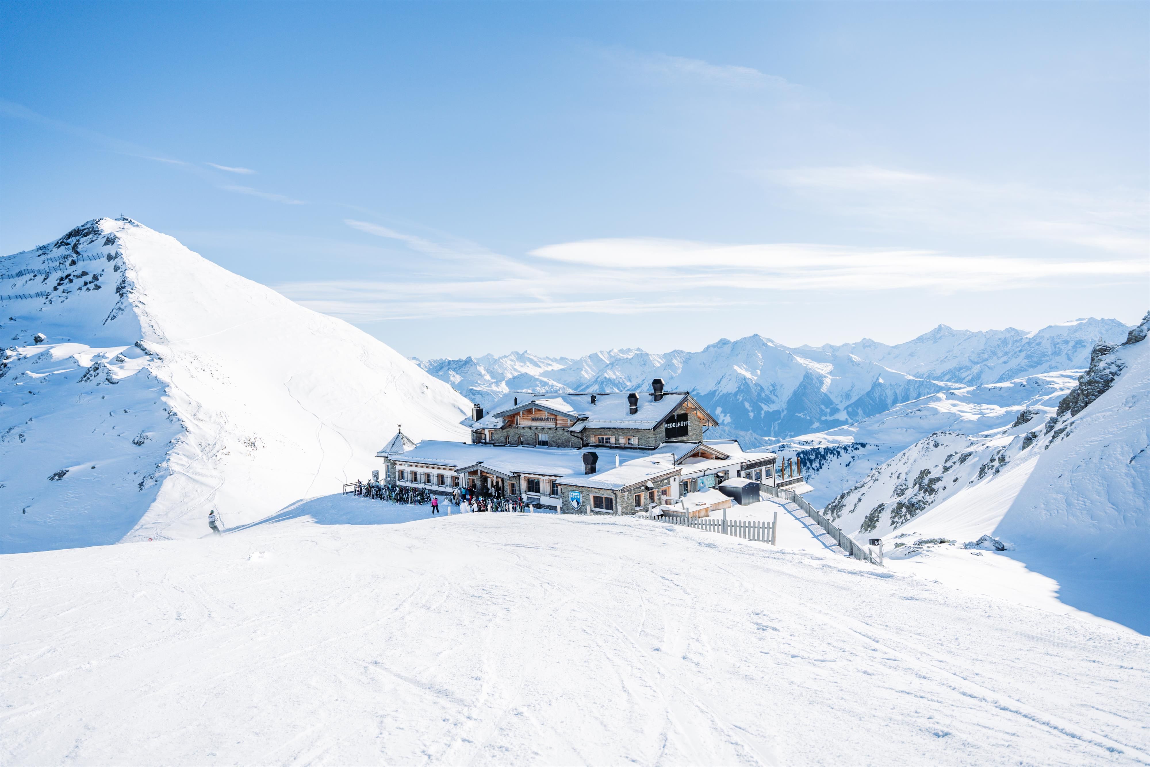 A mountain restaurant in the Alps, surrounded by snow-capped peaks. The sky is clear and bright blue.