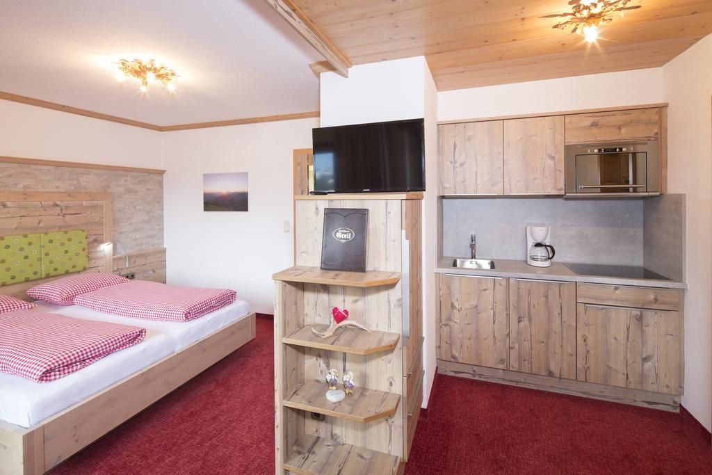 A cozy room with wooden decor, a double bed, and a small kitchenette. The room is bright and inviting with red carpet flooring.