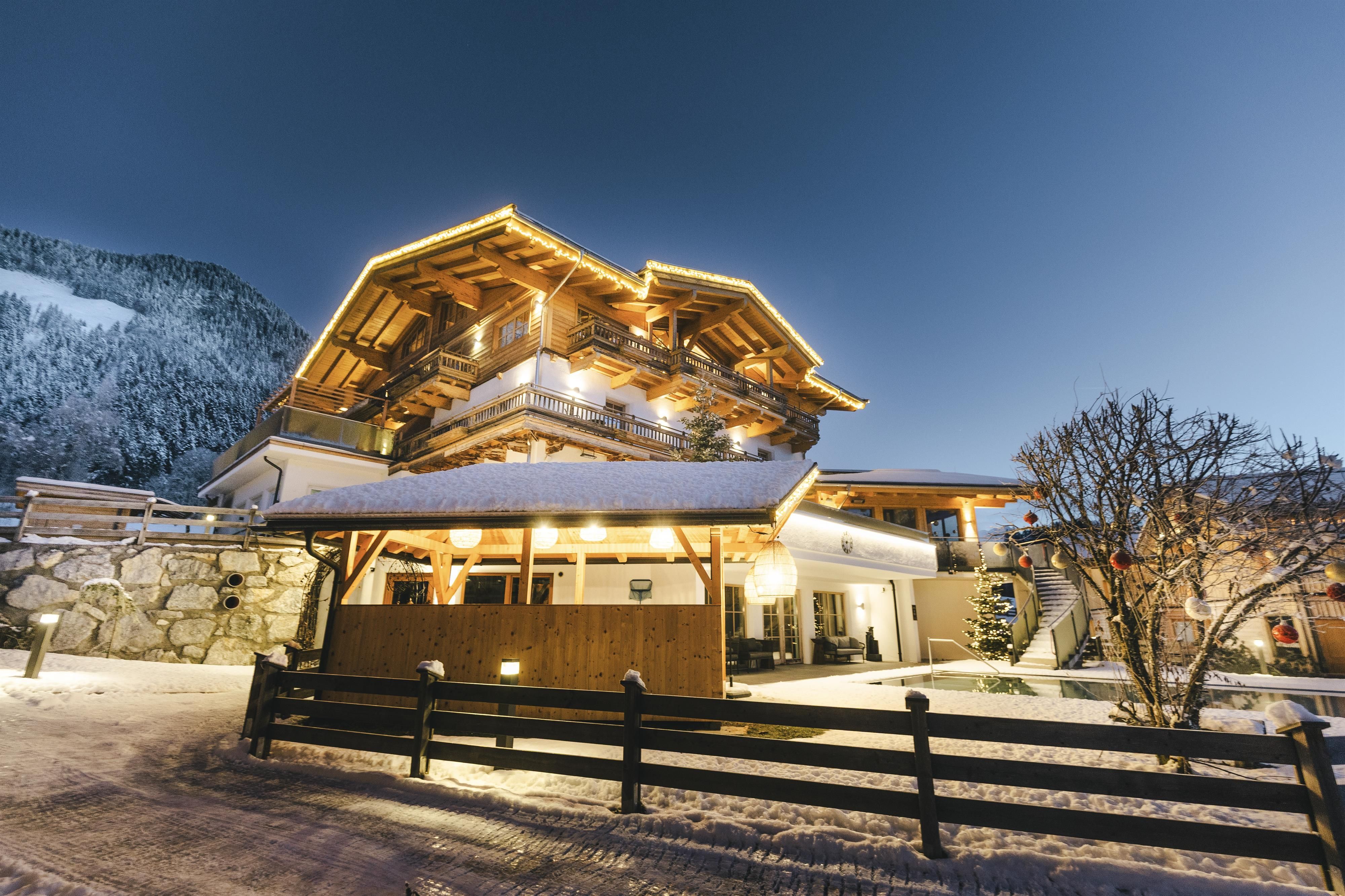 A charming mountain house in the snow, illuminated by warm light. Snow-covered mountains are visible in the background.