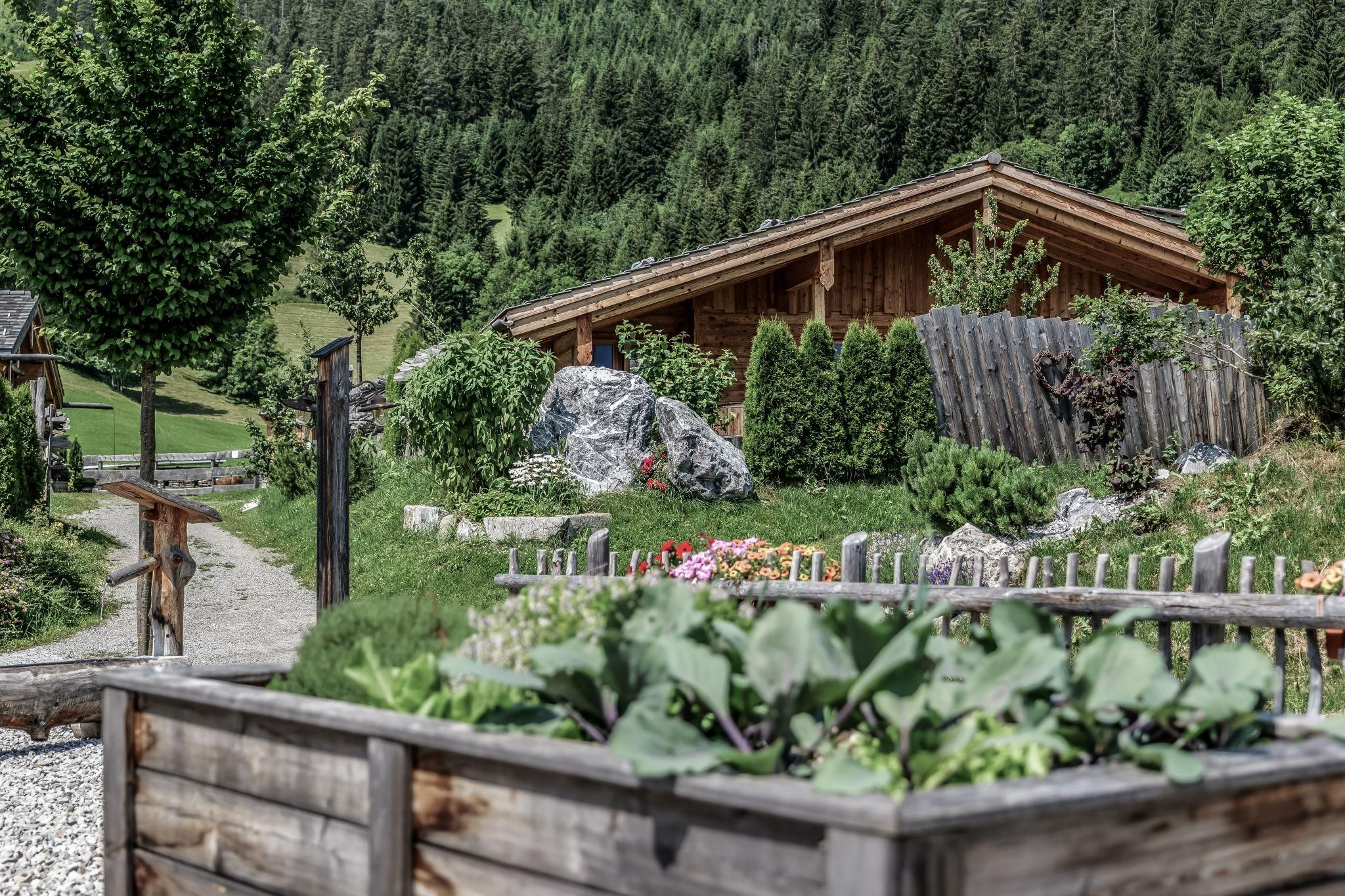 A cozy chalet surrounded by green landscape and trees. In the foreground, flowers and a vegetable garden can be seen.