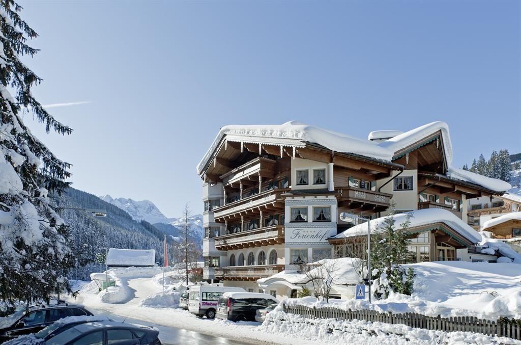 A charming hotel in the snow with multiple balconies and a traditional wooden design. The surroundings feature snow-covered mountains and a clear blue sky.