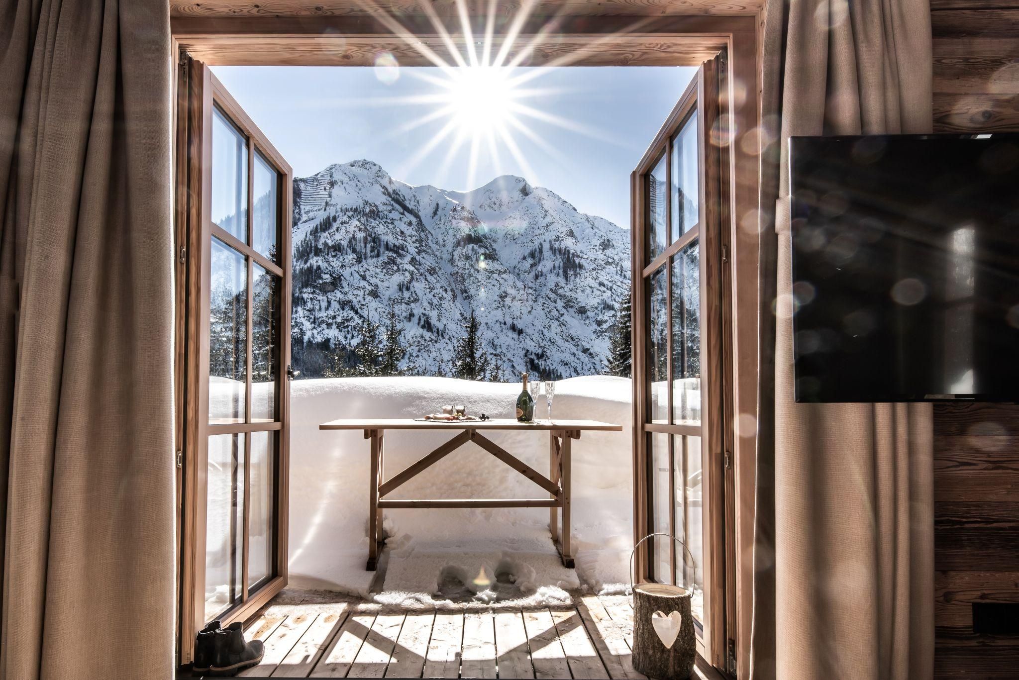 A view from a wooden window of impressive snow-covered mountains and radiant sunshine. Outside, there is a rustic table with a bottle.