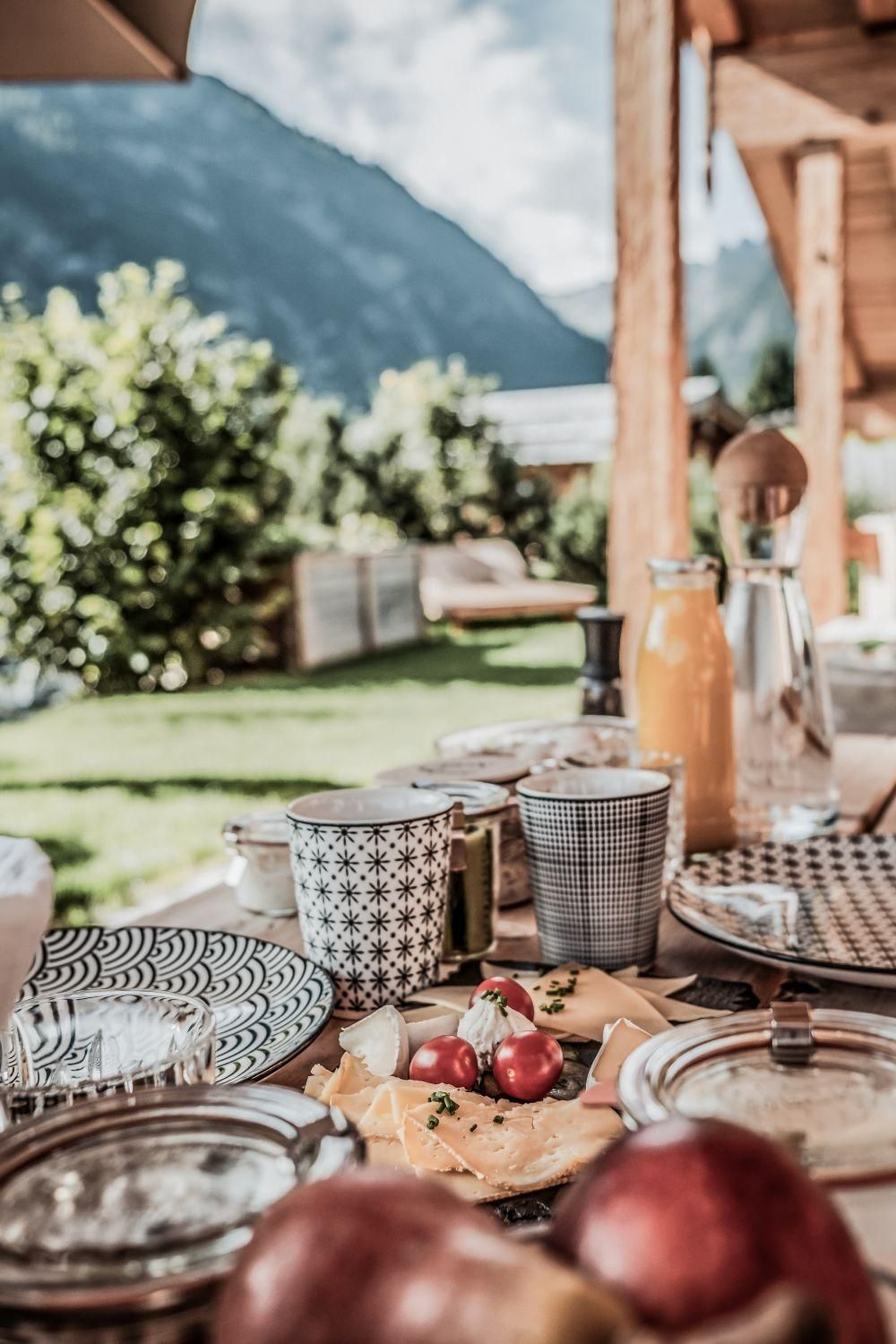 A beautifully set table outdoors with fresh fruit, cheese, and drinks. In the background, green trees and mountains can be seen.