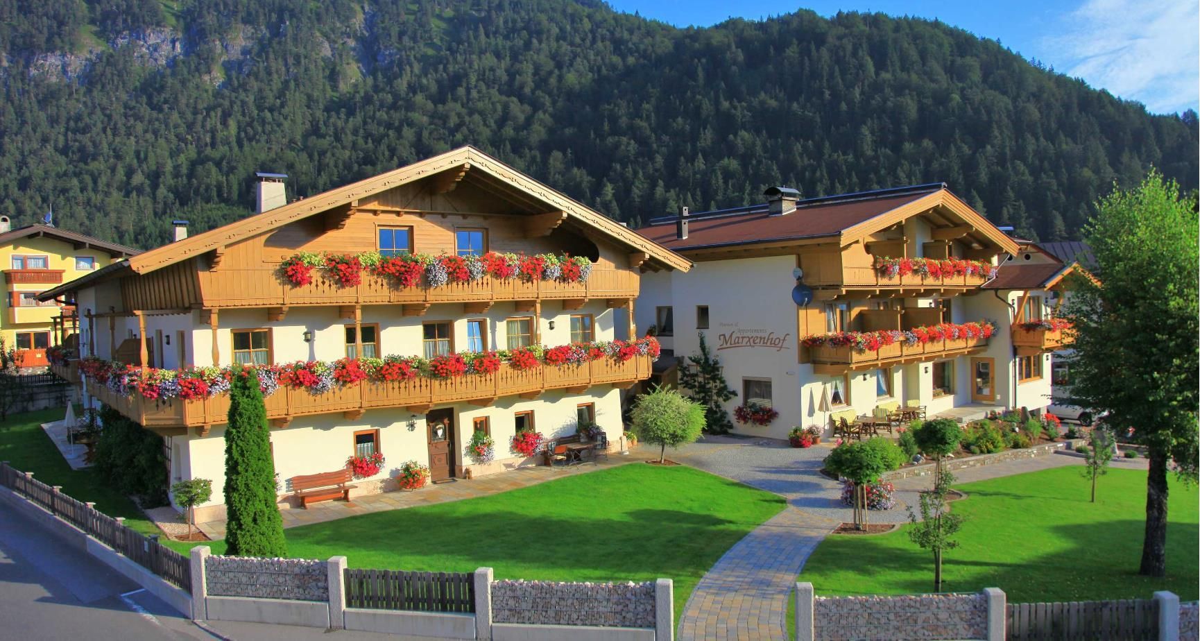 Two cozy buildings with balconies full of flowers surrounded by a well-tended garden. In the background, green hills and a clear sky can be seen.