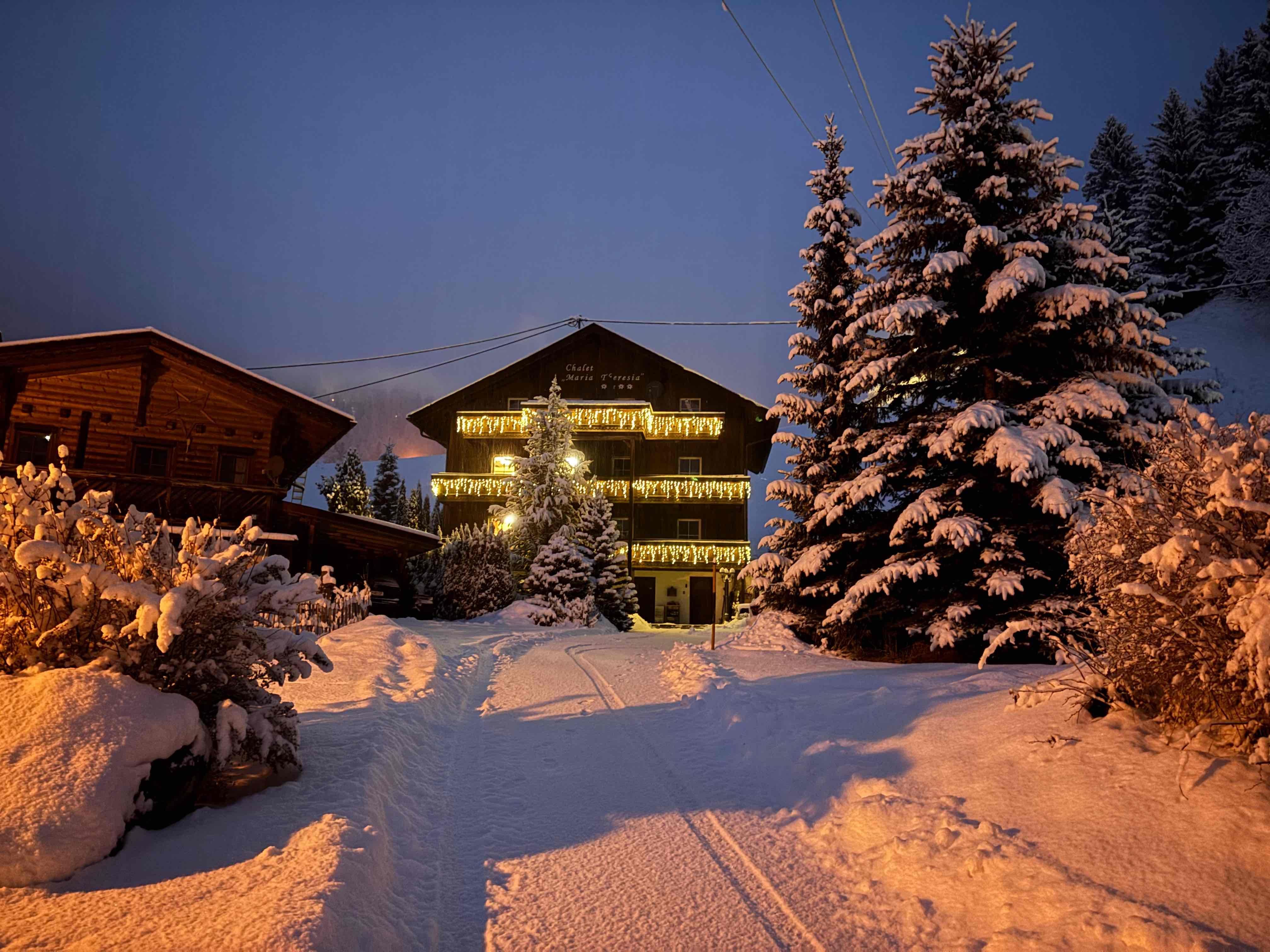 A cozy wooden house in the snow, surrounded by snow-covered fir trees. The dusk brings a warm light to the winter landscape.